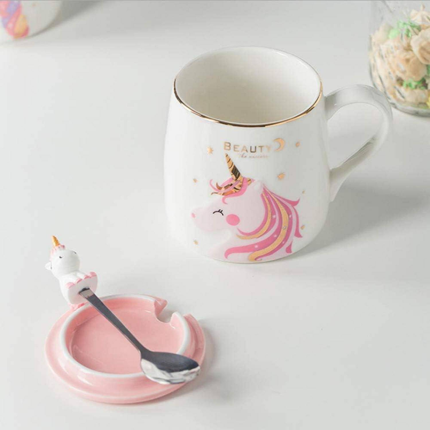 Weiße und rosa Einhorn-Tasse und Löffel auf einer weißen Oberfläche, mit 'Beauty'-Schriftzug auf der Tasse. Ein passendes Untersetzer hält den Löffel.