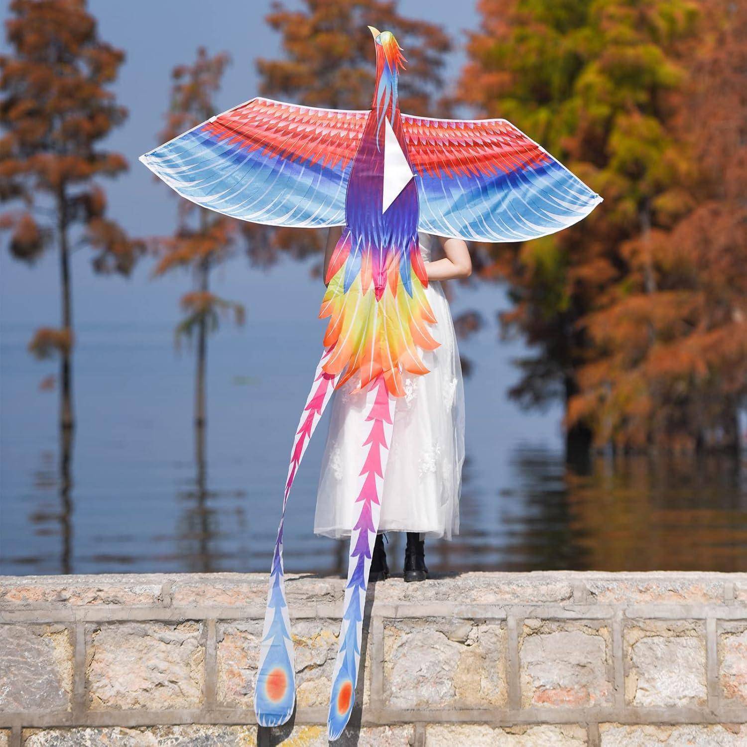 Eine Person in einem weißen Kleid steht auf einem Steinvorsprung und hält einen bunten, großen phönixförmigen Drachen, während im Hintergrund ein ruhiger See und Bäume zu sehen sind.