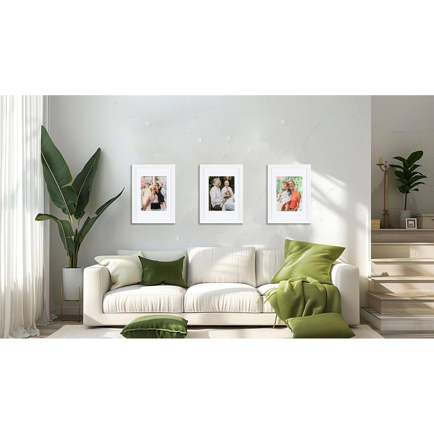 Ein beige Sofa mit grünen Kissen steht unter drei gerahmten Fotos an der Wand. Große Pflanzen befinden sich links und rechts und verleihen dem Raum eine natürliche Note.