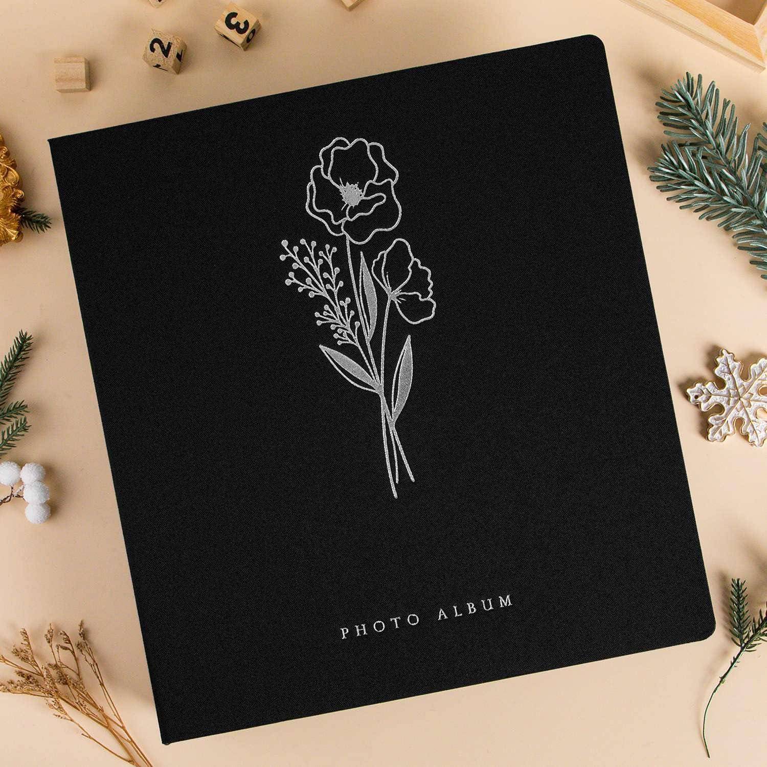 Schwarzes Fotoalbum mit einem geprägten Blumendesign auf dem Einband, umgeben von festlichen Dekorationen einschließlich Tannenzweigen und Weihnachtsschmuck.