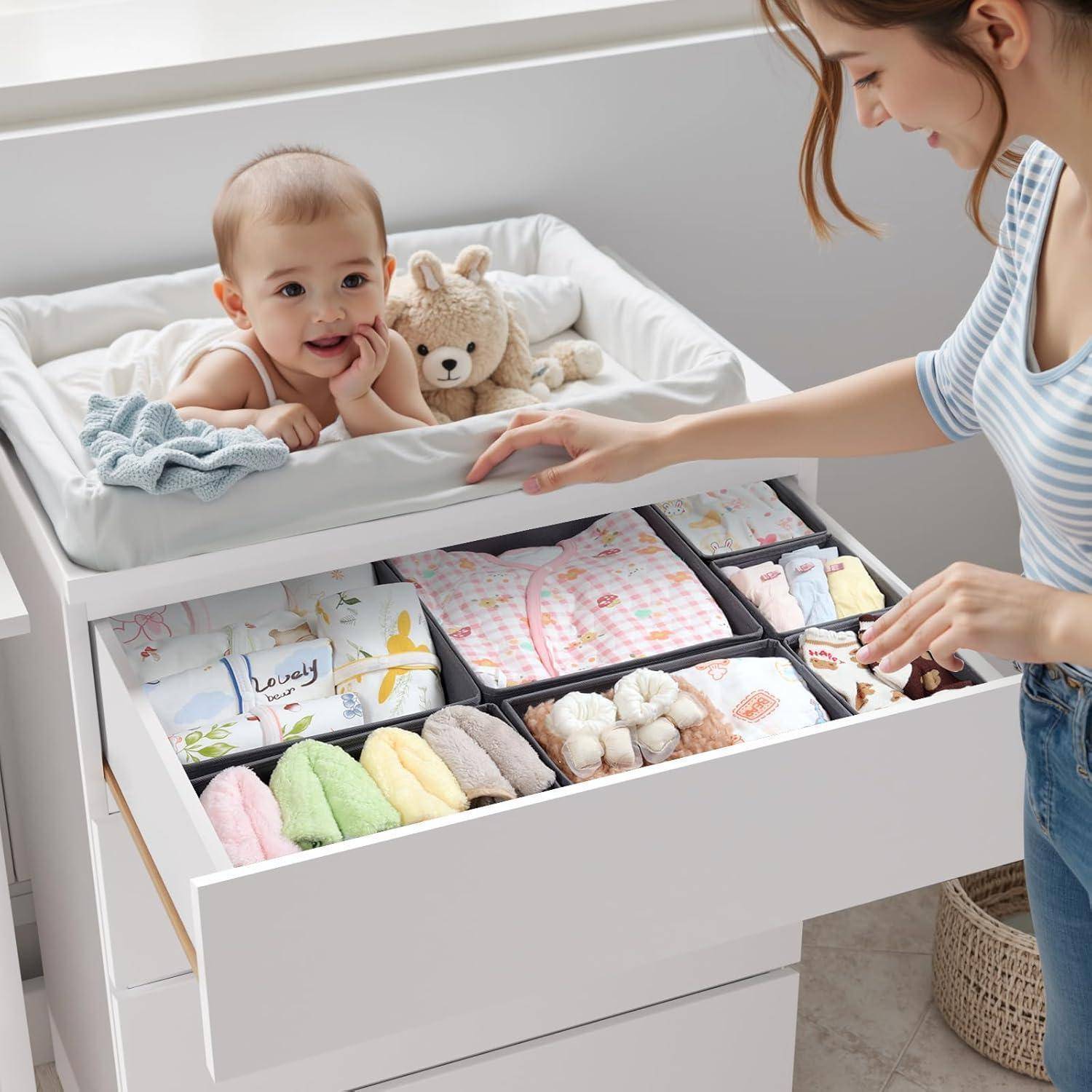 Eine Frau ordnet Babykleidung in einer Schublade an, während ein Baby auf einer Wickelkommode in der Nähe liegt und lächelnd ein Stofftier hält.