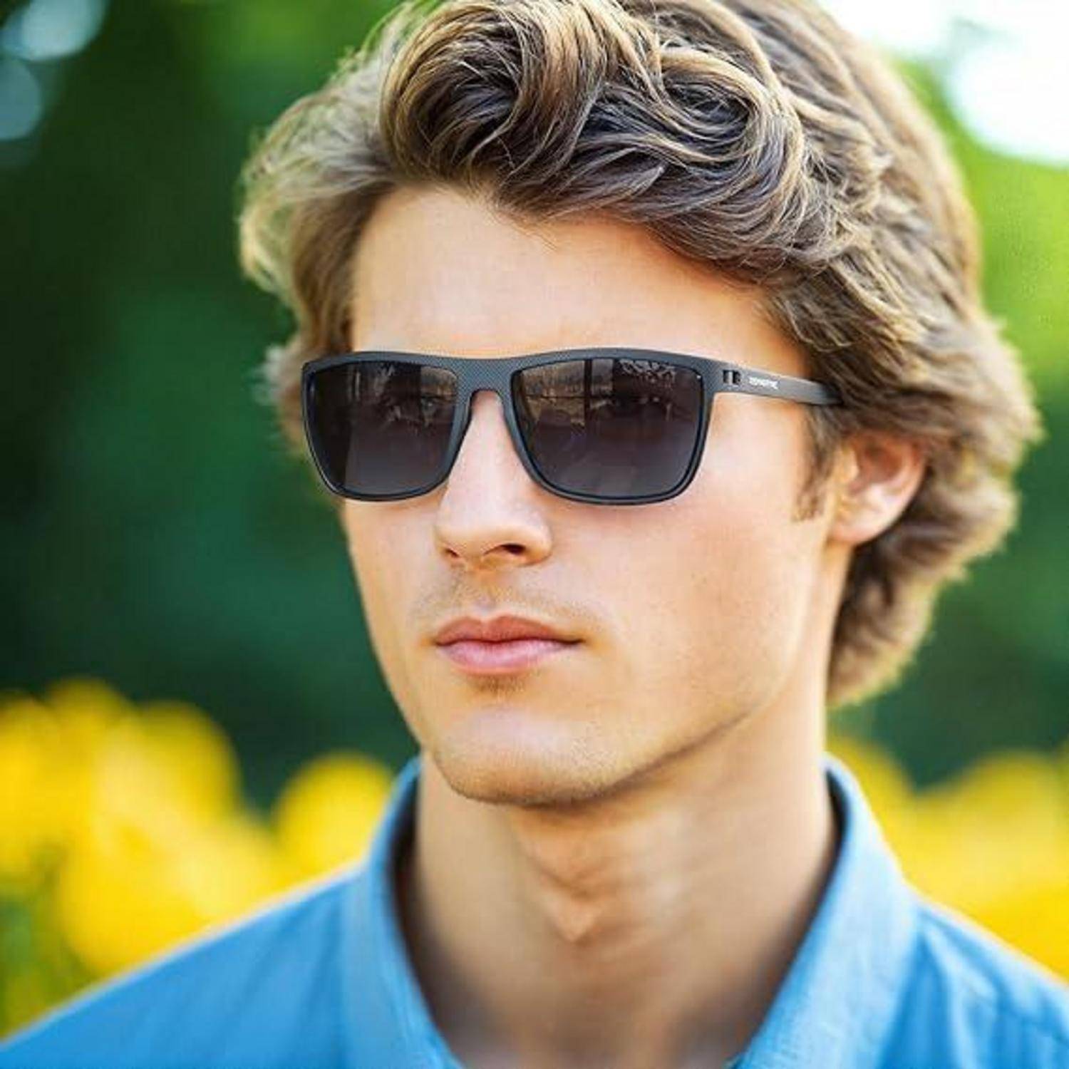 Eine Person mit Sonnenbrille steht im Freien; verschwommenes Grün und Blumen im Hintergrund deuten auf einen sonnigen Tag hin.