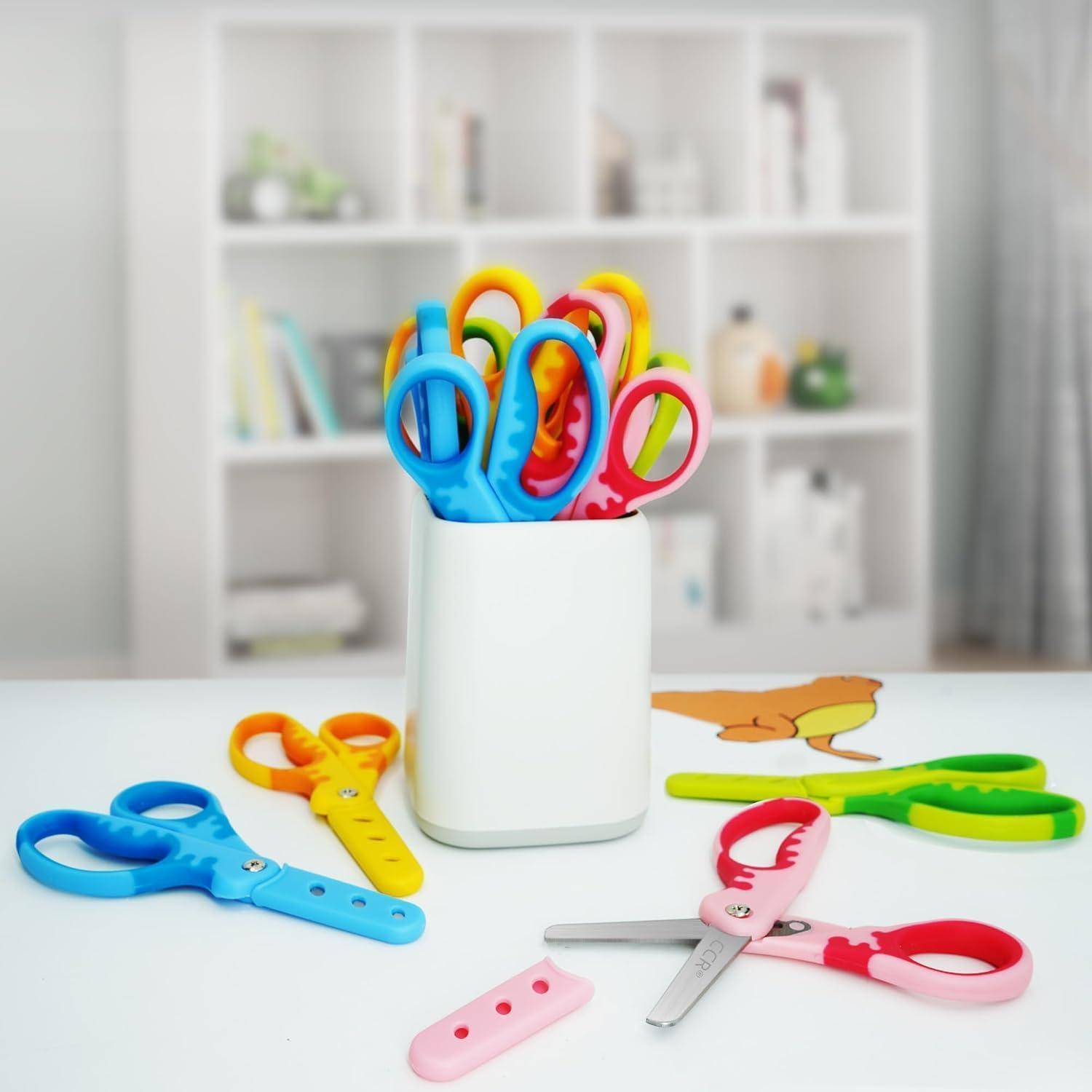 Eine Vielzahl bunter Kinderscheren in einem weißen Halter auf einem Tisch; Regale mit Büchern und Dekoration sind im Hintergrund unscharf.