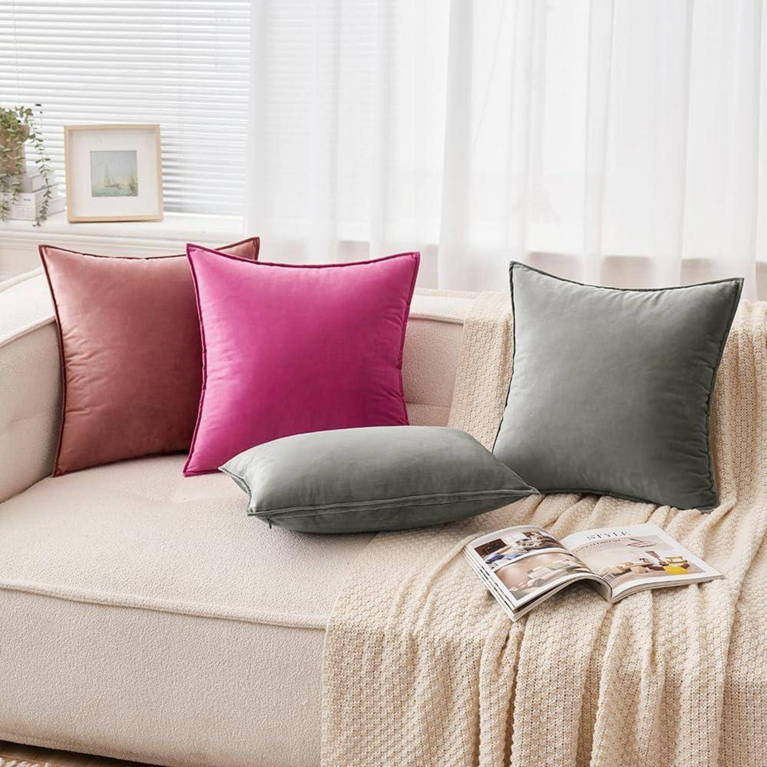 Ein beiger Sofa mit rosa, roten, grauen und grünen Kissen. Eine beige Decke mit einer Zeitschrift liegt auf der Armlehne. Eine kleine Pflanze und ein gerahmtes Bild stehen auf der Fensterbank hinter dem Sofa.