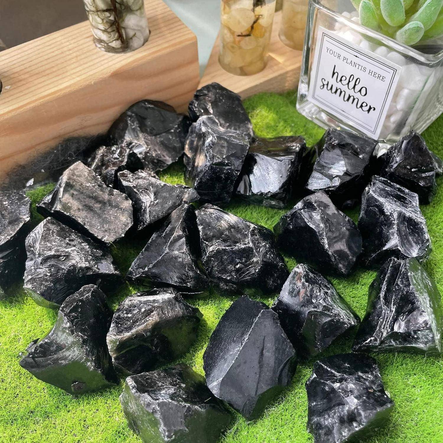 Schwarze Steine sind auf einer grünen strukturierten Oberfläche mit einem Holzrand angeordnet, mit kleinen Pflanzgefäßen im Hintergrund, wobei eines mit 