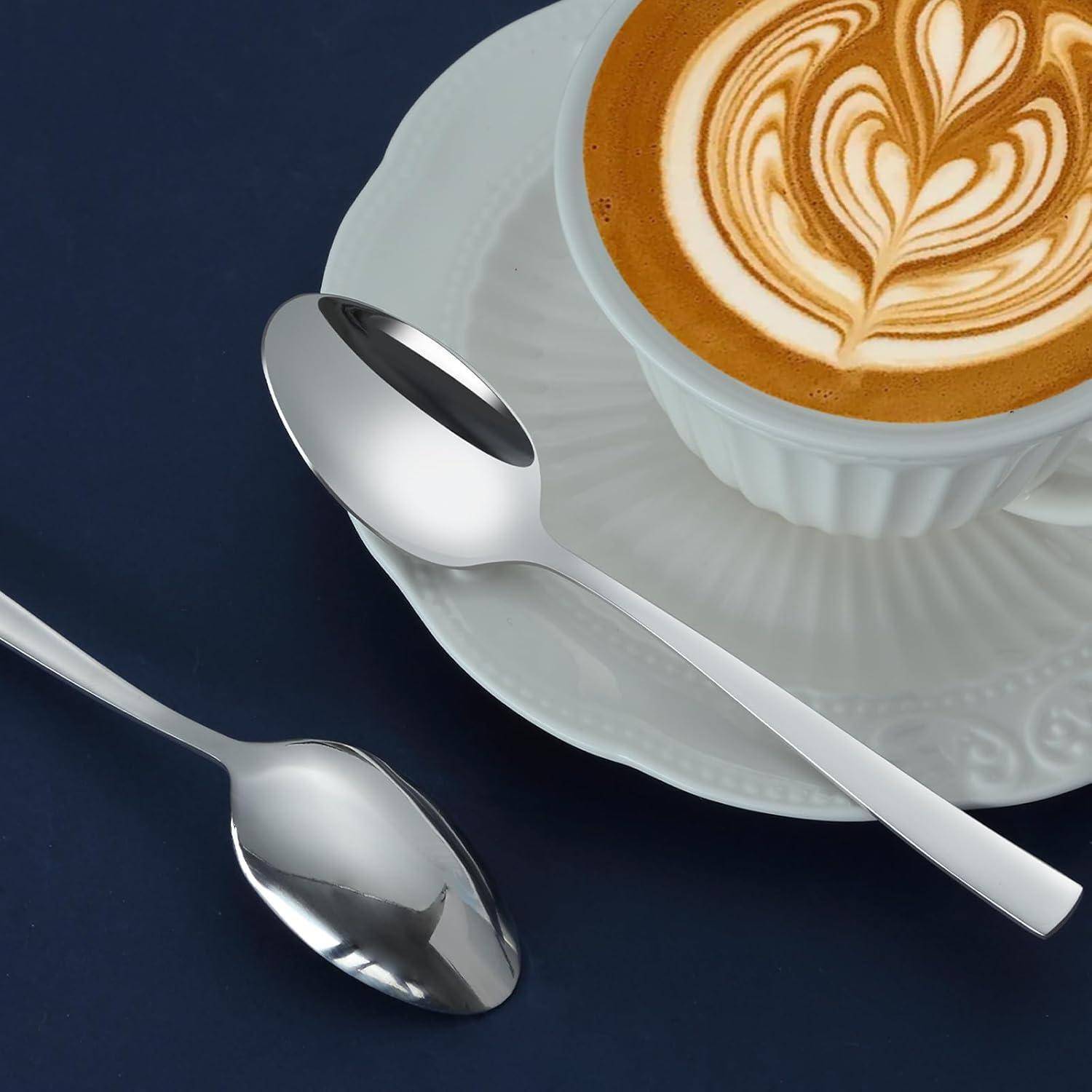 Ein Cappuccino mit Blatt-Latte-Art in einer weißen Tasse auf einem Unterteller, platziert auf einer dunklen Oberfläche neben zwei silbernen Löffeln.