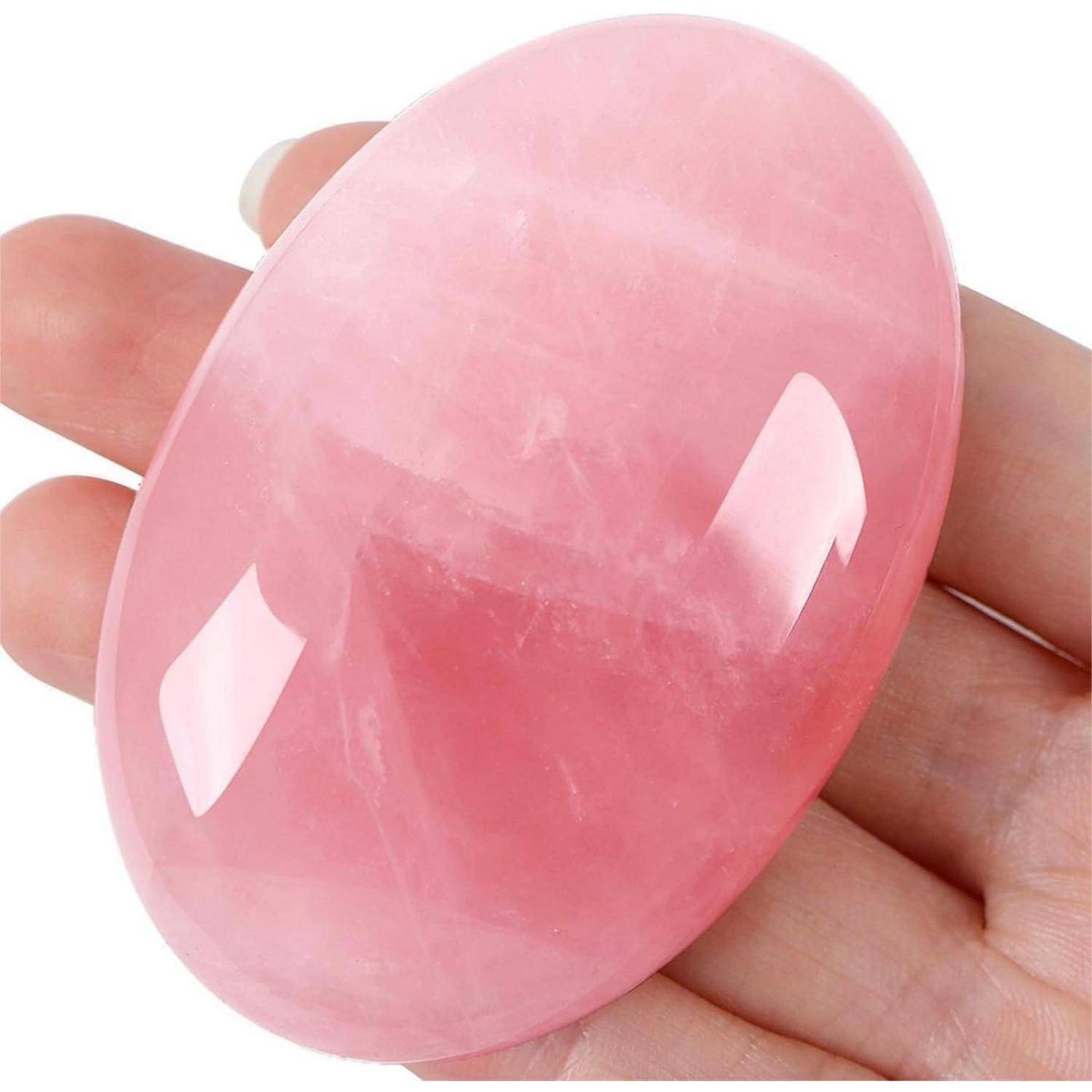 Eine Hand hält einen polierten Rosenquarz-Stein und zeigt seine glatte, ovale Form und rosafarbene Nuance, während er das Licht auf seiner Oberfläche reflektiert.
