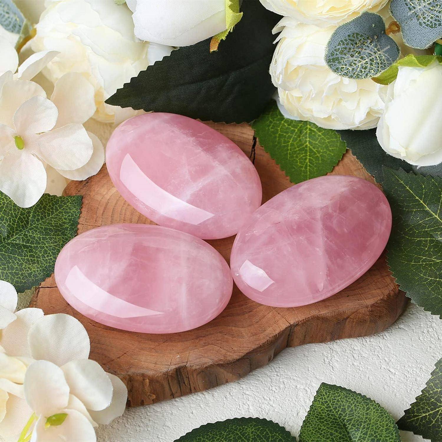 Drei polierte Rosenquarzsteine ruhen auf einer Holzbasis, umgeben von weißen Blumen und grünen Blättern, die eine beruhigende und natürliche Szenerie schaffen.