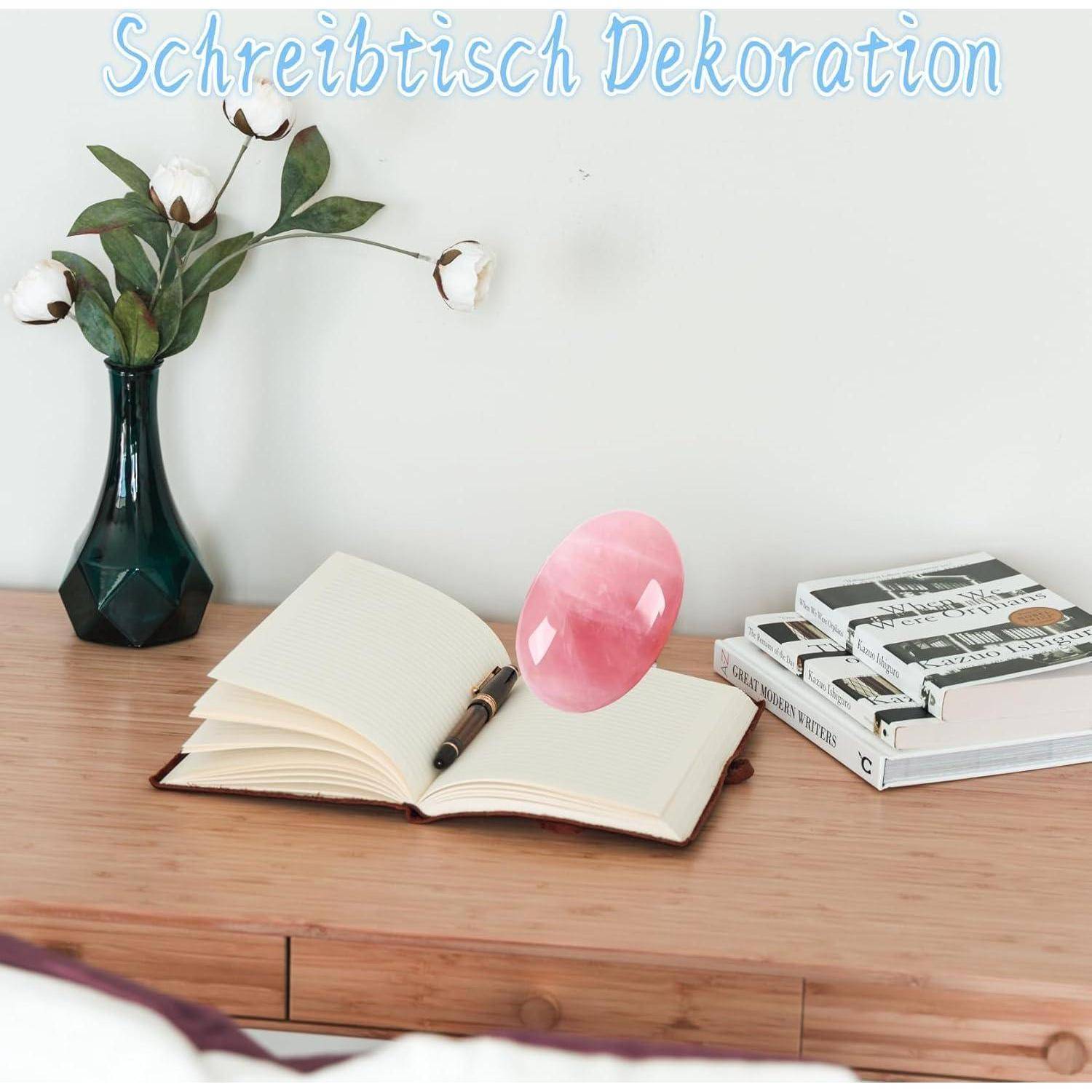 Ein Schreibtisch mit einem offenen Notizbuch, einem Stift, einer rosa Glasverzierung, einem Stapel Bücher, einer kleinen Vase mit Blumen und dem Text ‚Schreibtisch Dekoration' an der Wand.