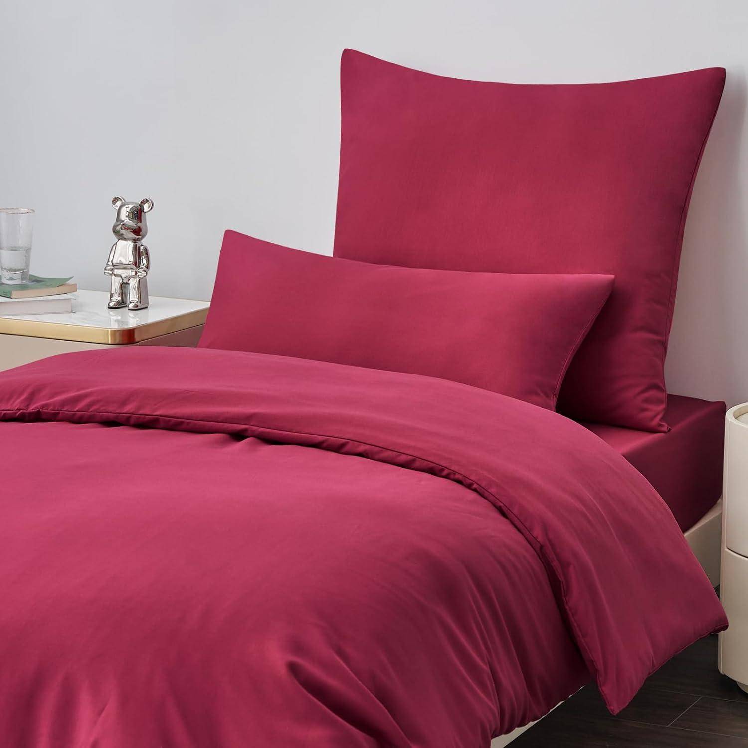 Ein ordentlich gemachtes Bett mit tiefroten Kissen und Bettdecke, daneben ein Nachttisch mit einer silbernen Teddybärfigur, einem Wasserglas und einem geschlossenen Buch.