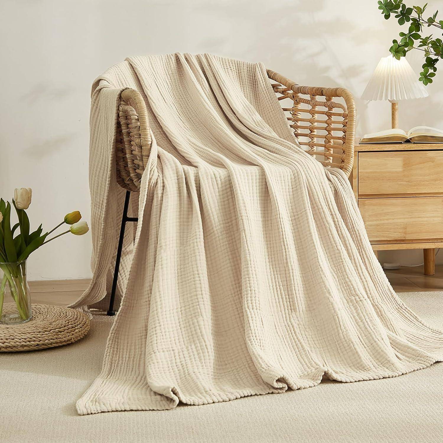 Eine gemütliche beige Decke, die über einem Korbstuhl neben einem kleinen Holztisch mit einer Lampe und einer Pflanze drapiert liegt und eine warme, einladende Atmosphäre schafft.