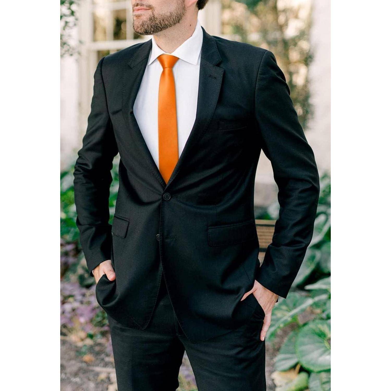Ein Mann in einem schwarzen Anzug mit orangefarbener Krawatte steht draußen, die Hände in den Taschen, vor einem unscharfen Gartenhintergrund.