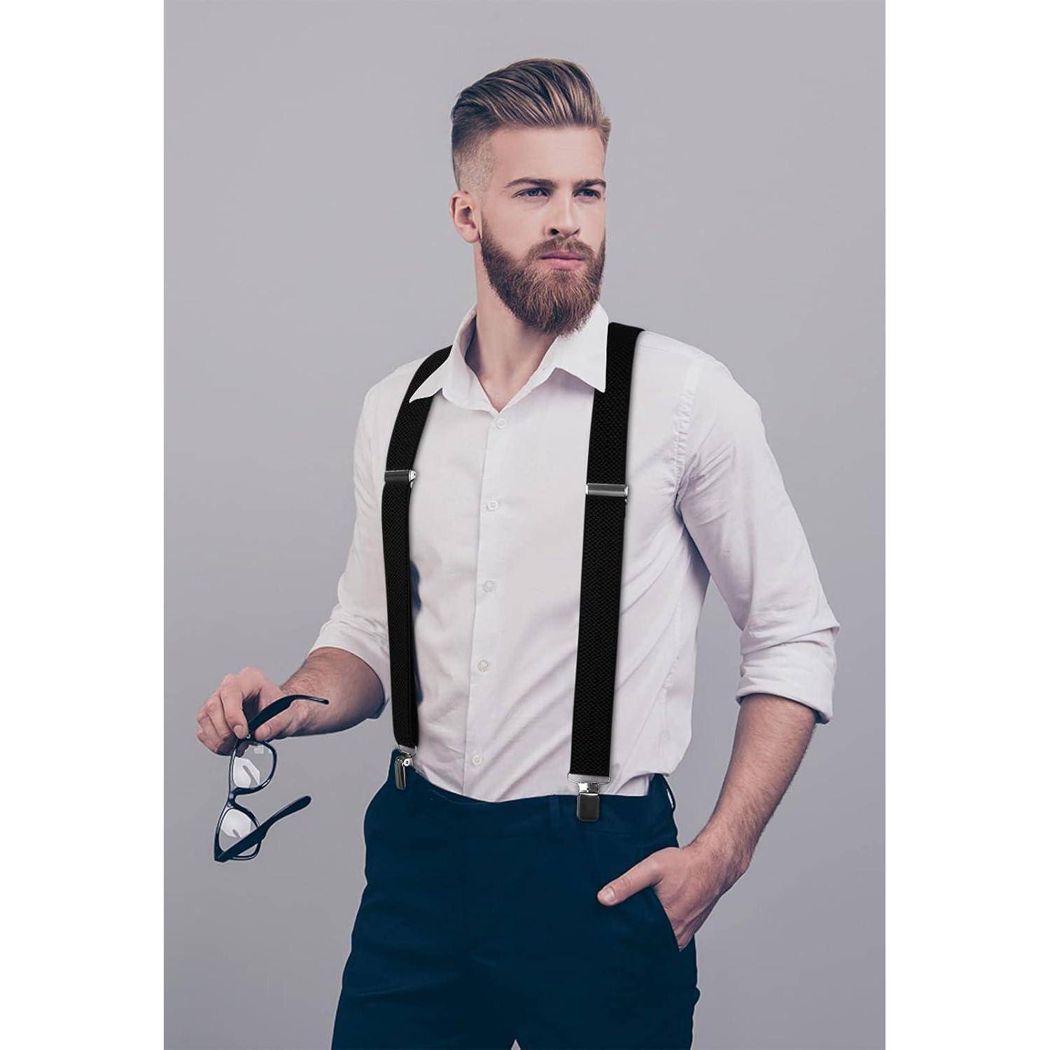 Ein Mann mit Bart, der ein weißes Hemd und schwarze Hosenträger trägt, hält eine Brille in einer Hand. Er blickt mit einem neutralen Gesichtsausdruck zur Seite.