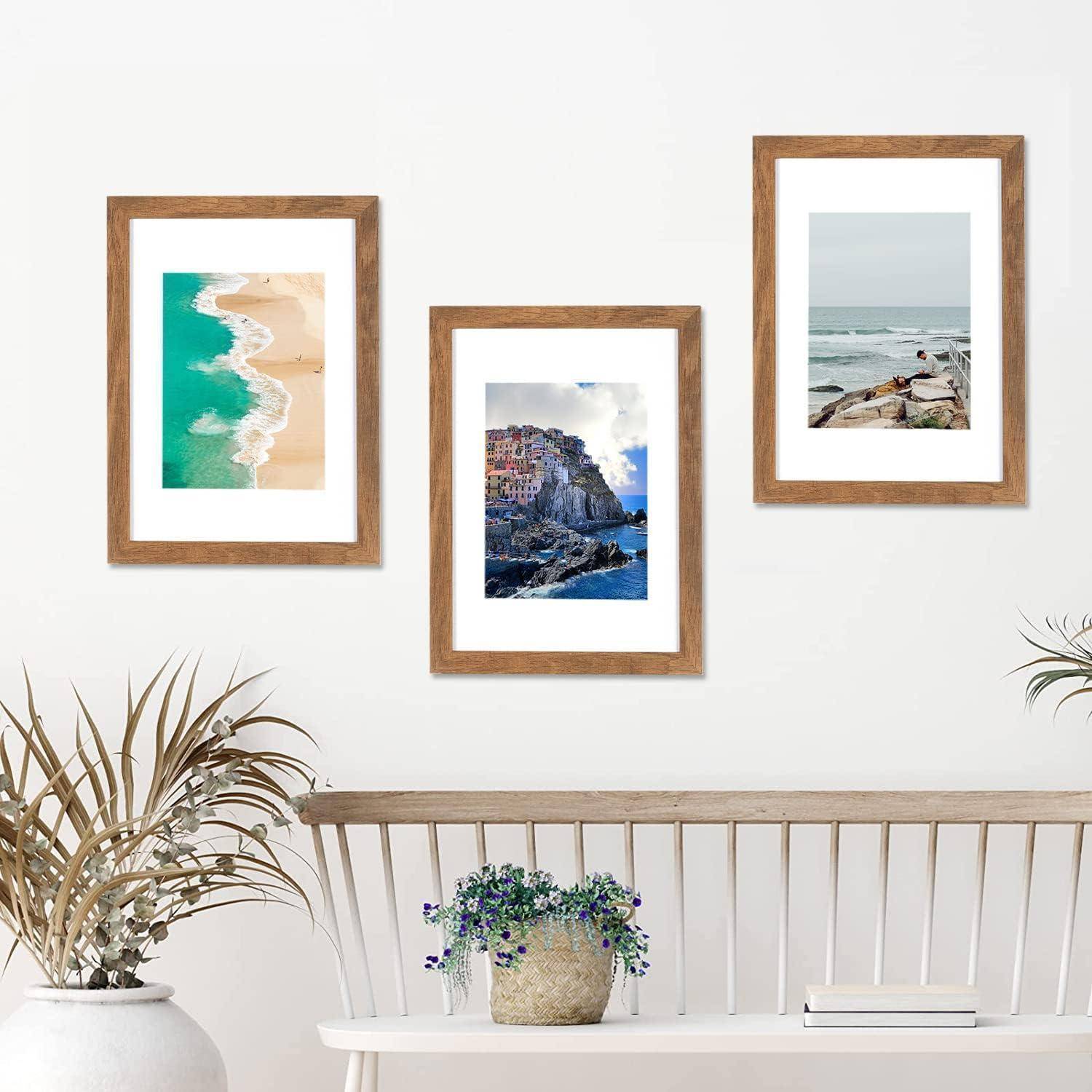 Drei gerahmte Fotos an einer weißen Wand über einer Bank: ein Strand, eine Küstenstadt auf einem Felsen und ein felsiger Küstenabschnitt. Die Dekoration umfasst eine Topfpflanze.