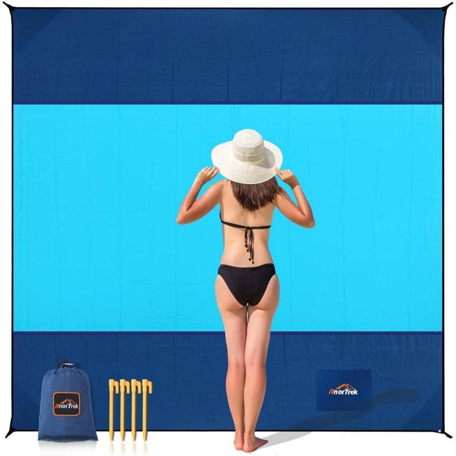 Eine Frau in einem schwarzen Bikini und Sonnenhut steht vor einem zweifarbigen blauen Strand-Zelt-Hintergrund. Gelbe Stangen und eine blaue Tasche liegen auf dem Sand.