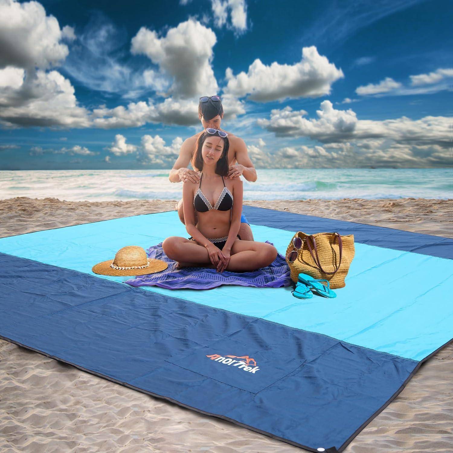 Ein Paar sitzt auf einer großen Stranddecke am Meer, wobei die Frau einen Hut trägt und eine Tasche neben sich hat. Der Himmel ist teilweise bewölkt.