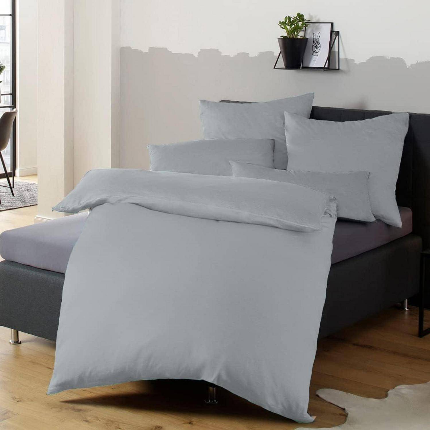 Ein sorgfältig gemachtes Bett mit hellgrauer Bettwäsche und mehreren Kissen ist in einem modernen Raum mit Holzböden und neutralen Wänden platziert.
