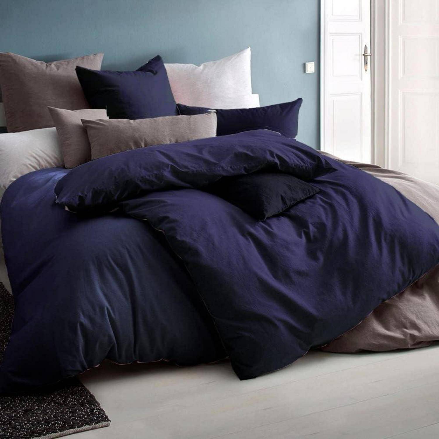 Ein Bett mit blauen und grauen Kissen und einer marineblau-farbenen Bettdecke in einem hellen Raum mit einer blauen Wand und einer weißen Tür im Hintergrund.