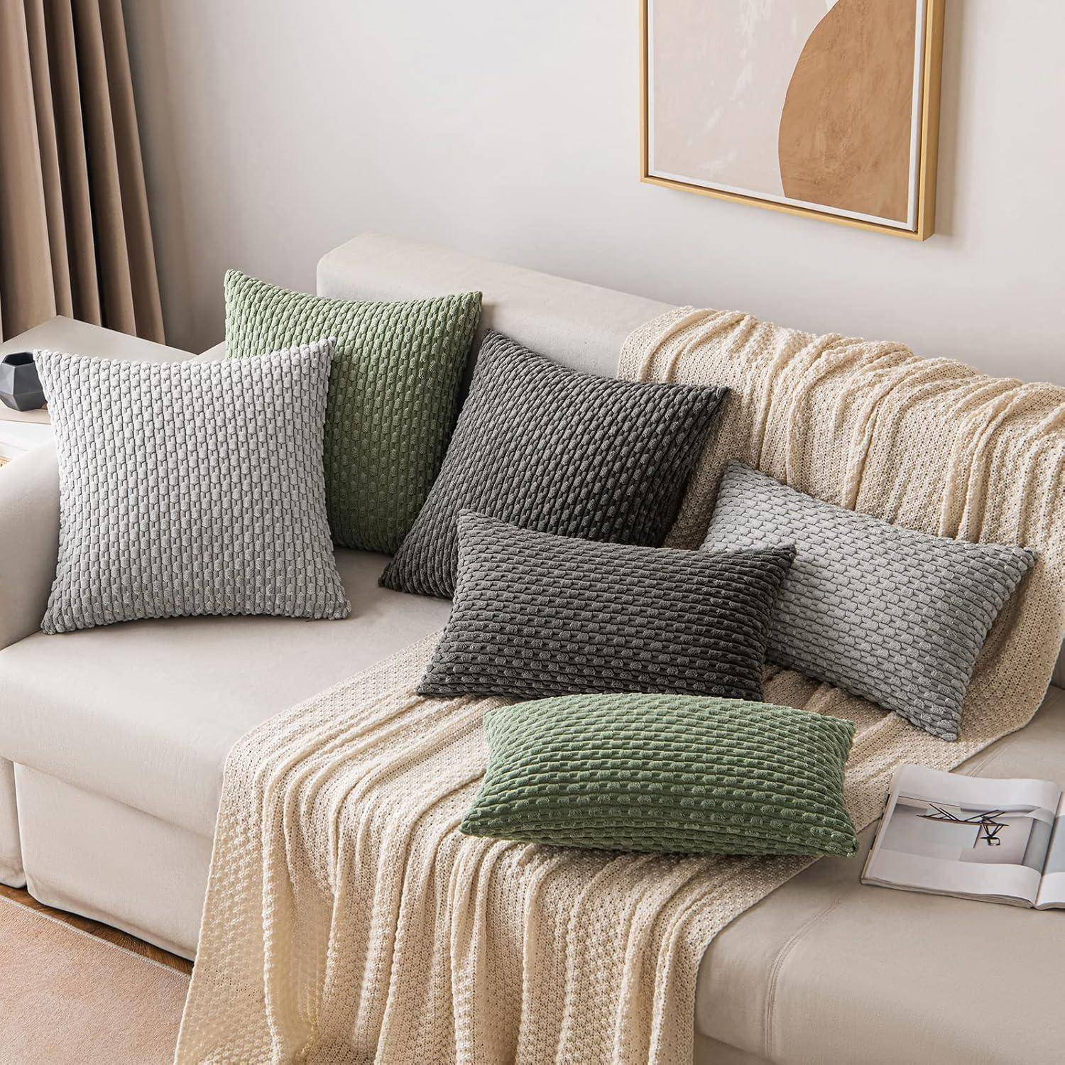 Eine gemütliche Wohnzimmerszene mit einem weißen Sofa, das mit cremefarbenen gestrickten Decken bedeckt ist und mit strukturierten grauen und grünen Kissen verziert wird. Ein minimalistisches gerahmtes Kunstwerk hängt darüber.