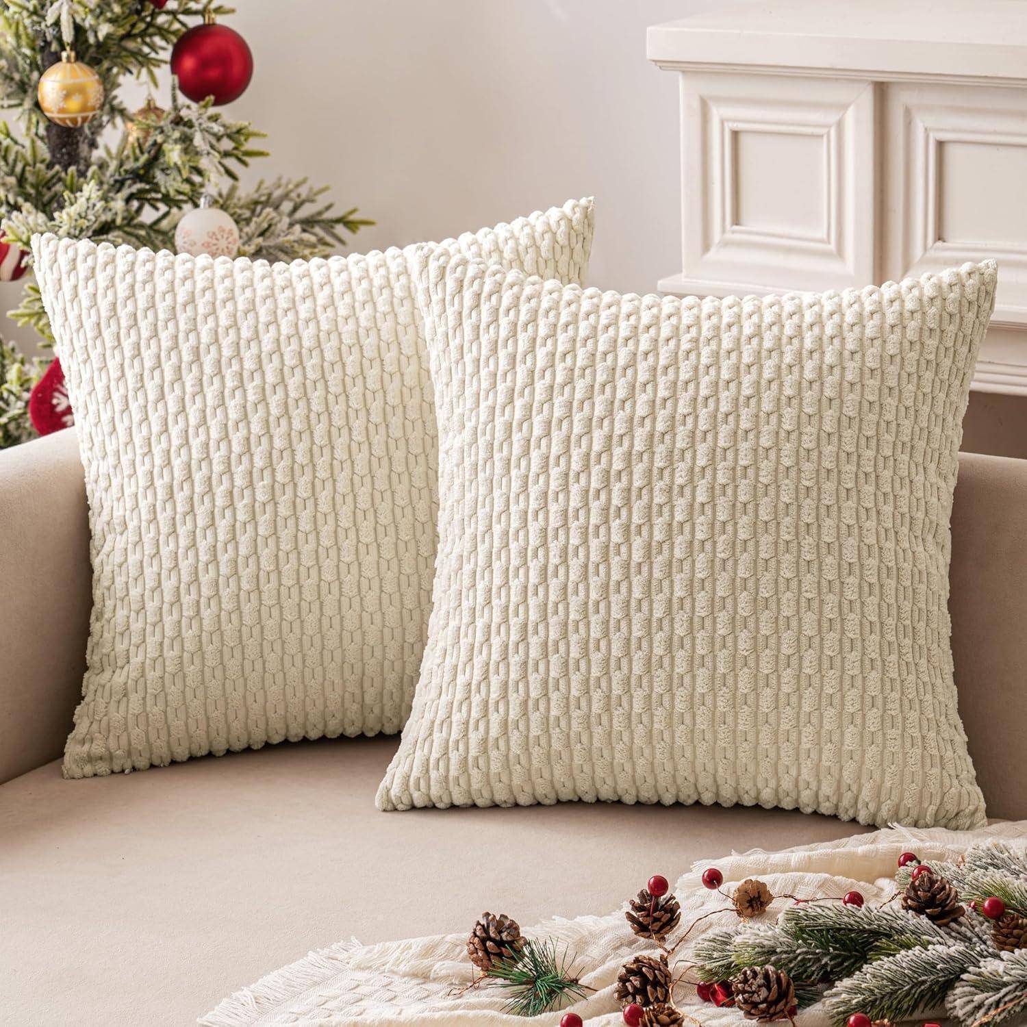 Zwei strukturierte weiße Kissen auf einem beigen Sofa, mit einer festlichen Tannzapfen-Anordnung im Vordergrund und einem teilweise sichtbaren geschmückten Baum.