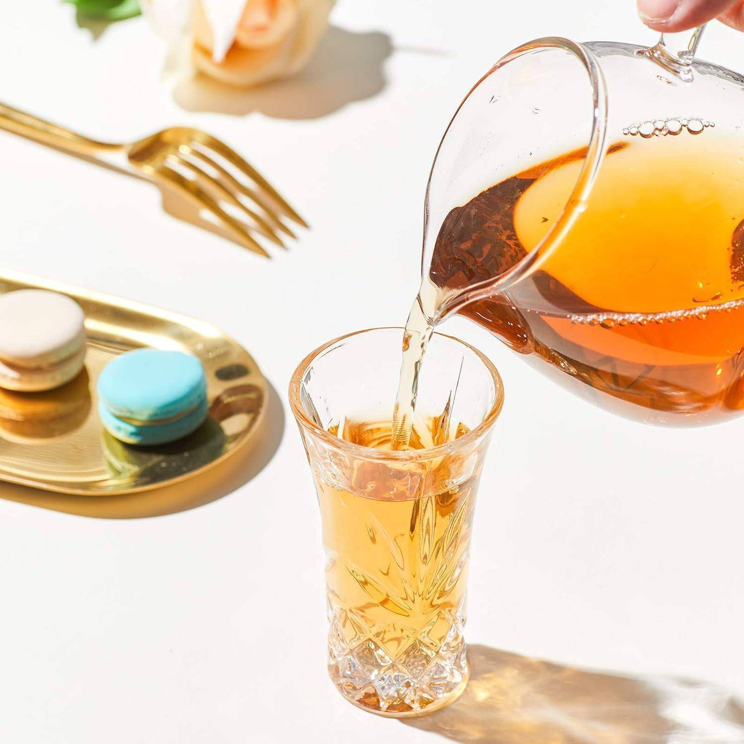 Eine Hand gießt Tee aus einer durchsichtigen Teekanne in ein Glas. In der Nähe liegen bunte Macarons auf einem goldenen Tablett mit einer Gabel und einer weißen Rose.