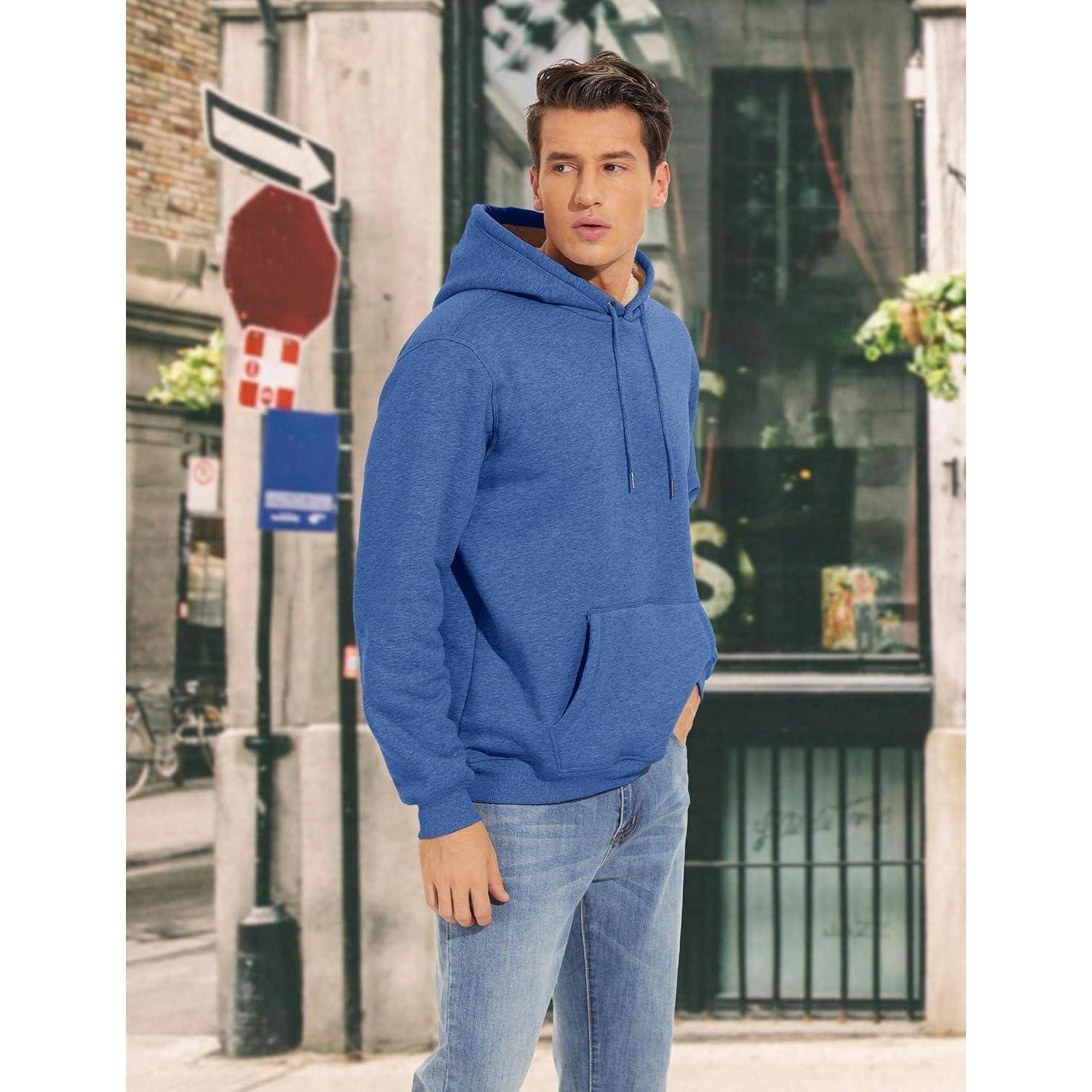 Eine Person in einem blauen Hoodie und Jeans steht auf einer Stadtstraße mit Gebäuden und Grünflächen im Hintergrund.
