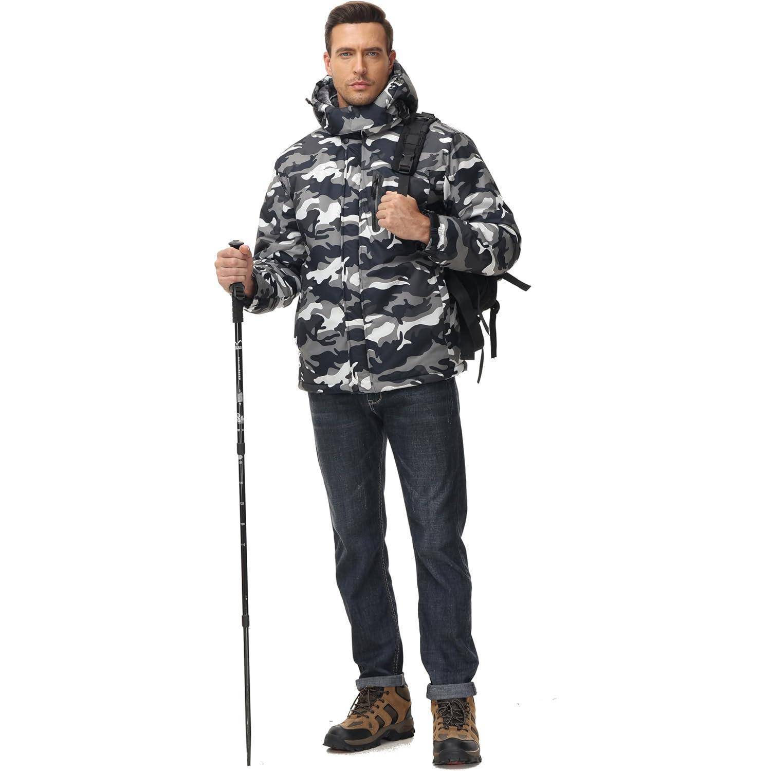 Ein Mann in einer grauen Tarnjacke und Jeans steht mit einem Rucksack und Wanderstock, was darauf hindeutet, dass er für Outdoor-Aktivitäten vorbereitet ist.