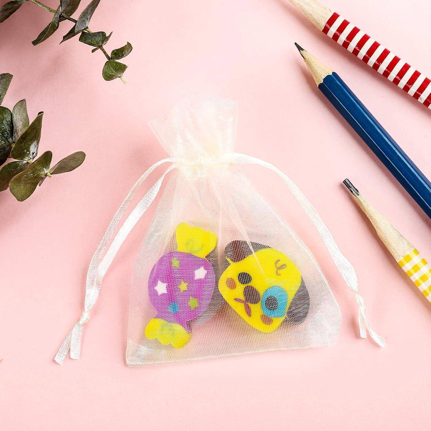 Kleine weiße Kordelzugtasche mit bunten Radiergummis in Form von Süßigkeiten und Pizza, die zwischen Bleistiften und Grünpflanzen auf einer rosa Oberfläche platziert ist.