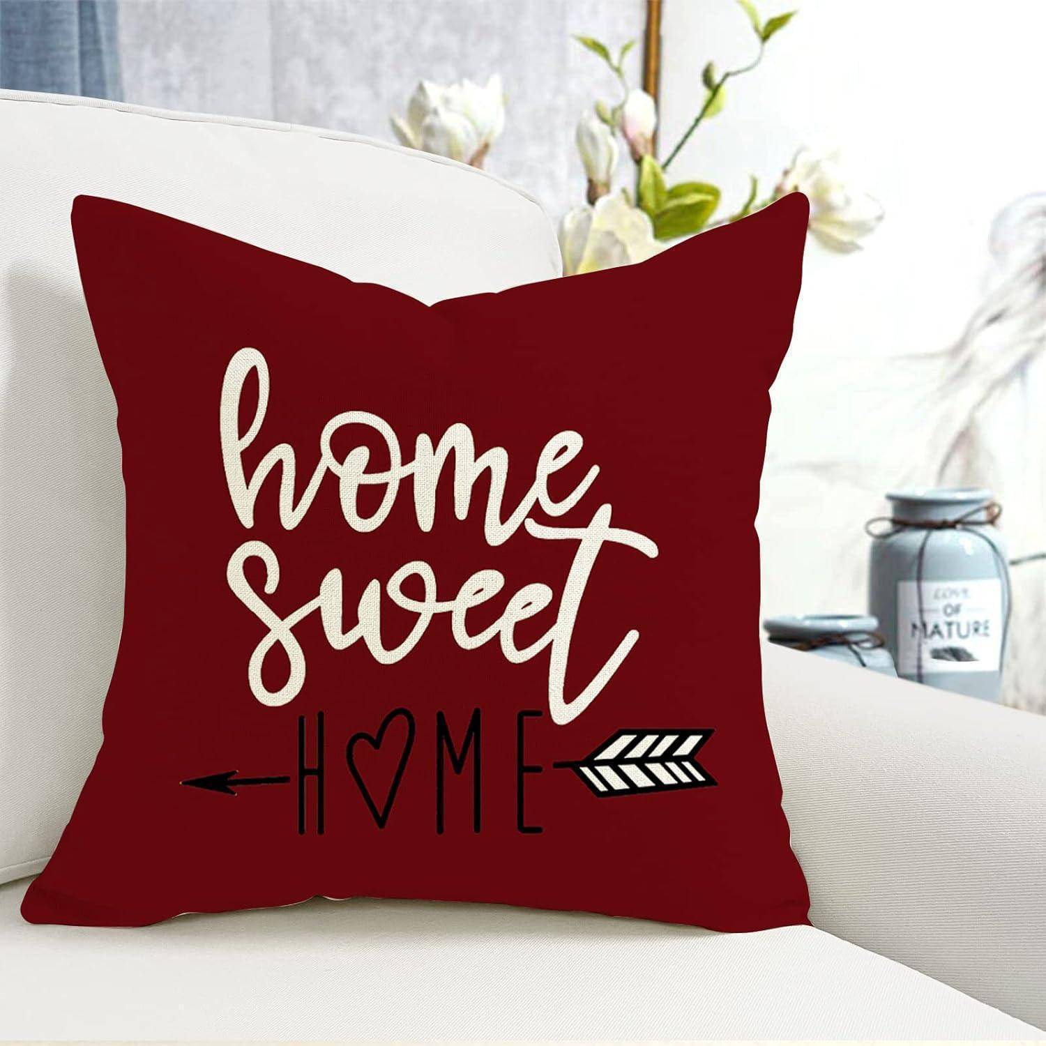 Ein rotes Zierkissen mit dem Text „home sweet home" in weißer Kursivschrift, versehen mit einem Pfeil und einem Herz unter dem Text. Blumen im Hintergrund.