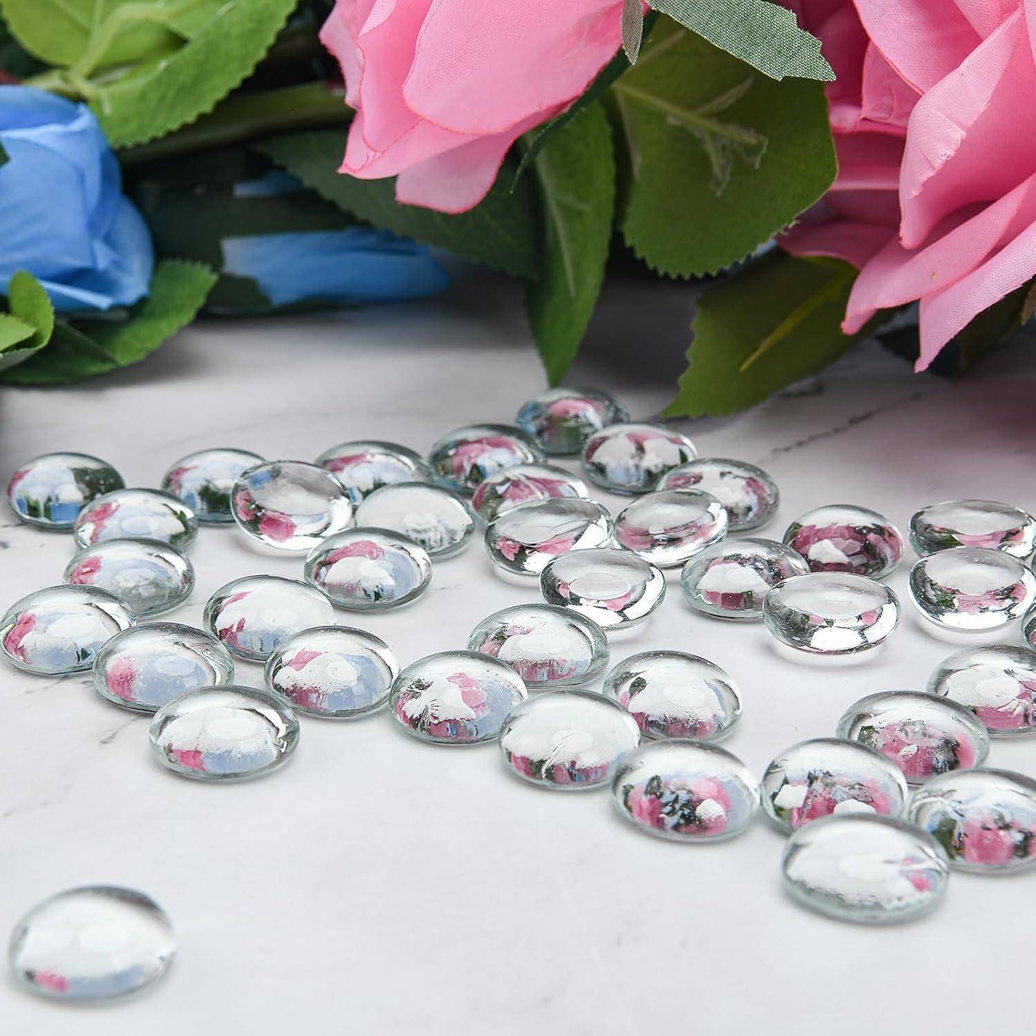 Klare Glaskiesel, verstreut auf einer Marmoroberfläche, umgeben von rosa und blauen künstlichen Blumen mit grünen Blättern.