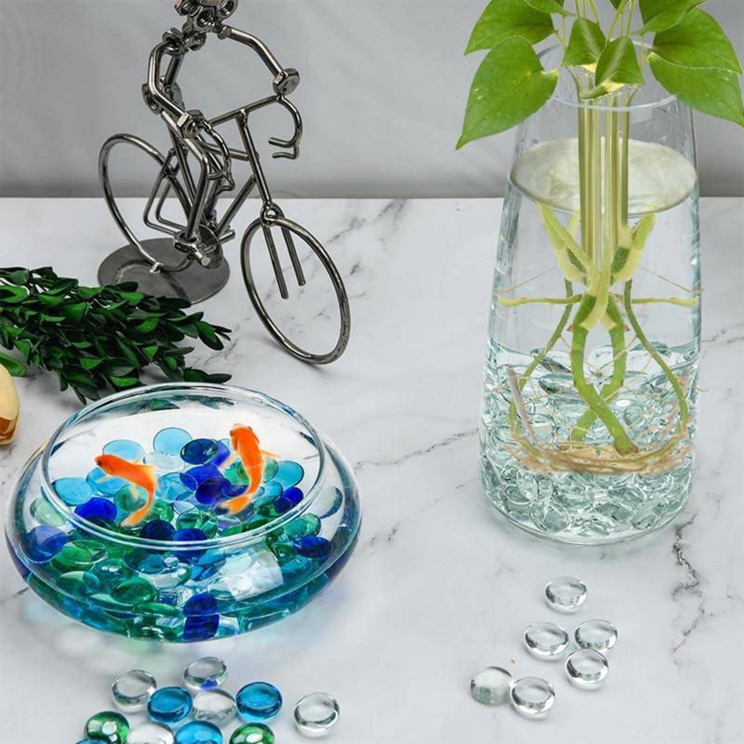 Ein dekoratives Stillleben mit einer Metallskulptur eines Radfahrers, einer runden Glasschale mit bunten Murmeln und einer Vase mit einer grünen Pflanze.