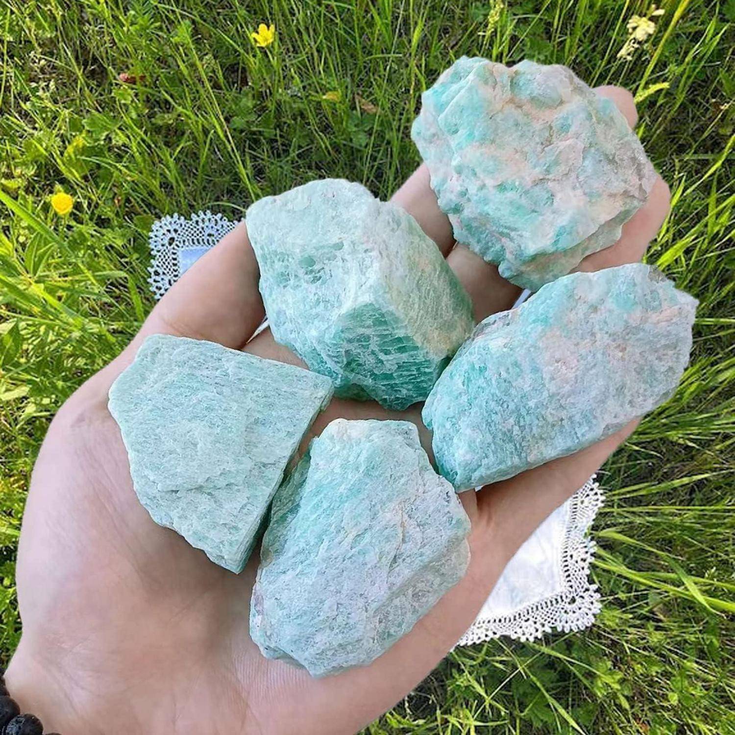 Eine Hand hält fünf grobe, pastellgrüne Steine vor einem Spitzendeckchen und grünem Gras im Hintergrund.