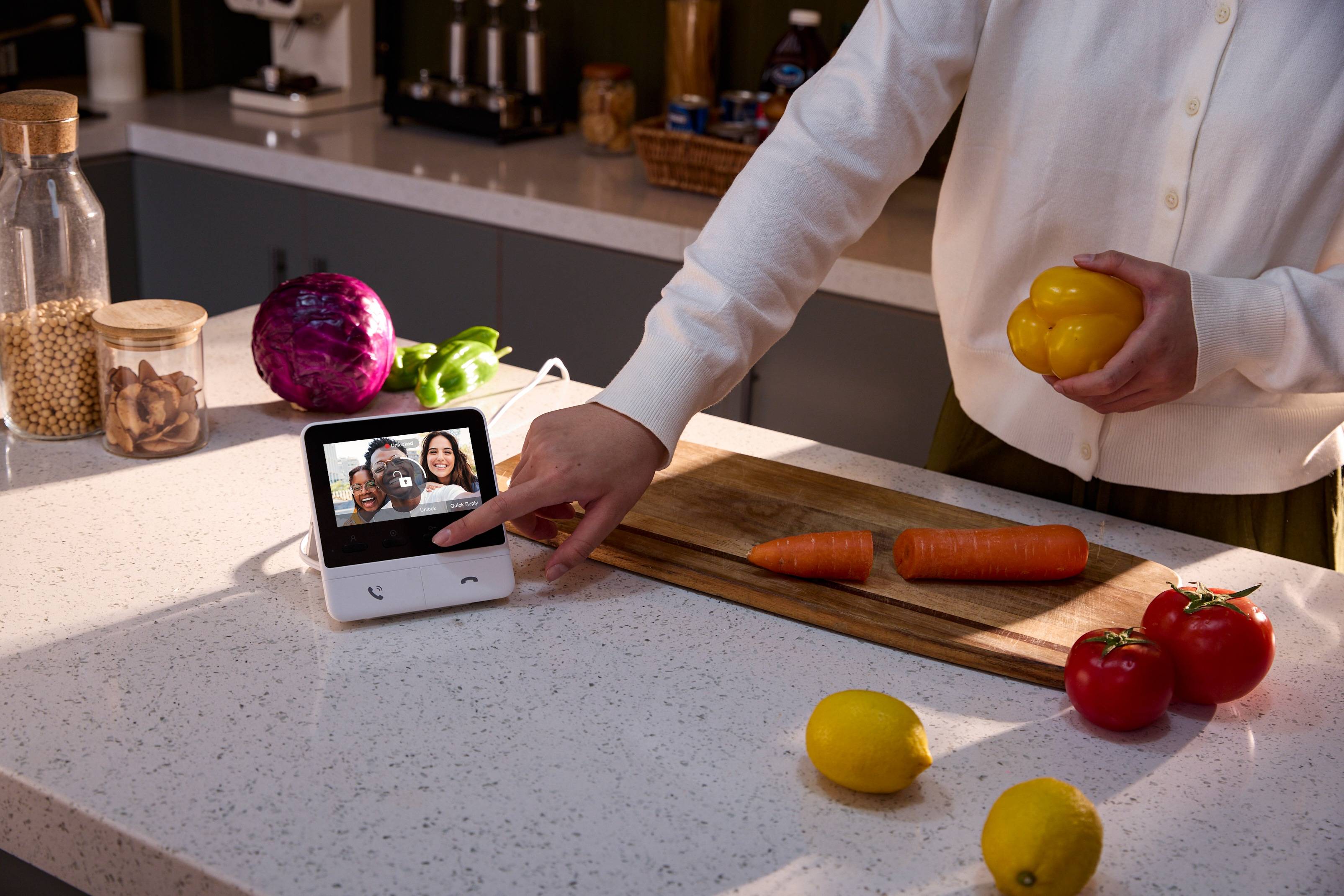 Eine Person in einer Küche interagiert mit einem Smart-Display, das einen Videoanruf mit zwei Personen zeigt. Gemüse und Obst liegen auf der Theke.