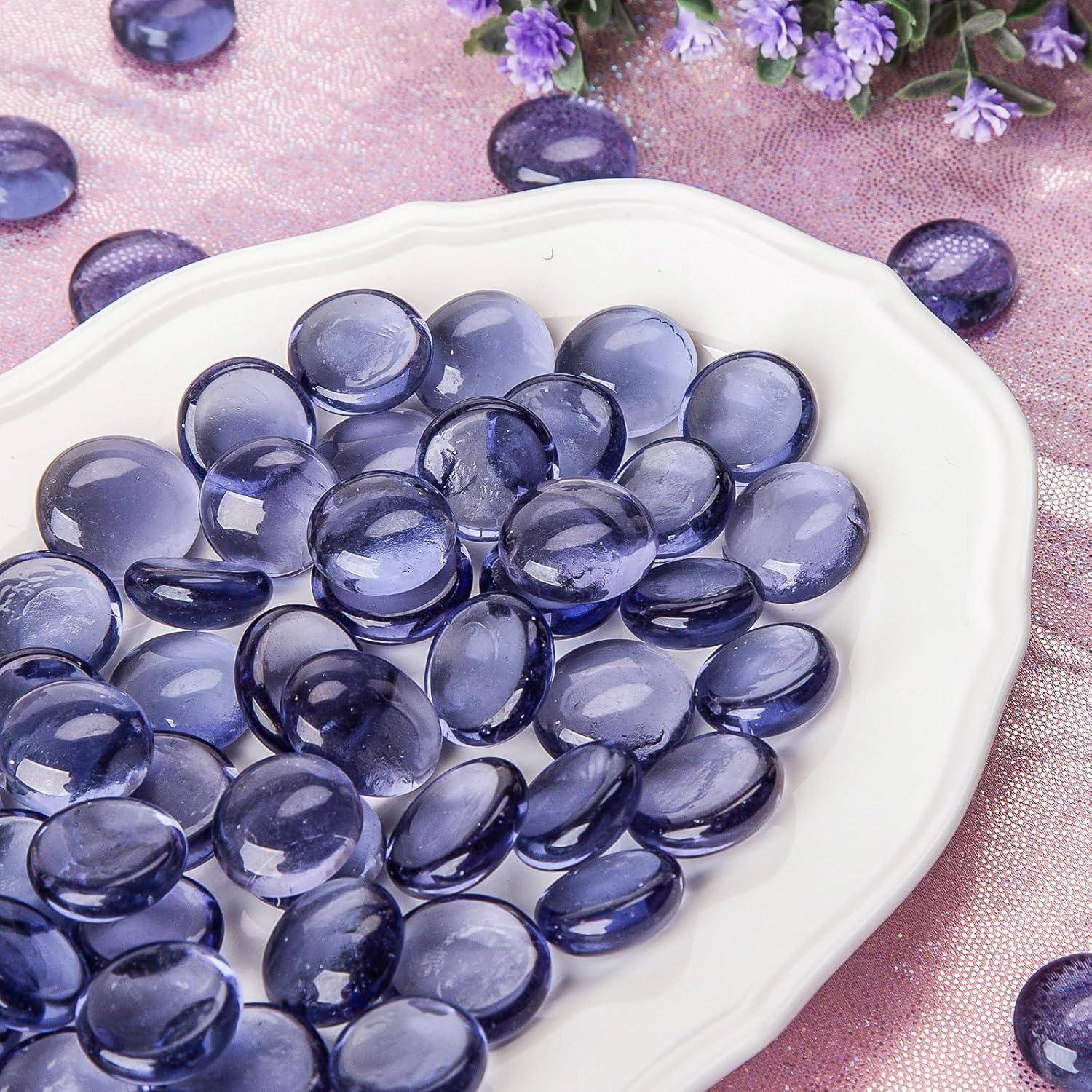Eine weiße Schale, gefüllt mit glatten, durchscheinenden blauen Glassteinen auf einem pinken Stoffhintergrund, umgeben von verstreuten Steinen und kleinen violetten Blumen.