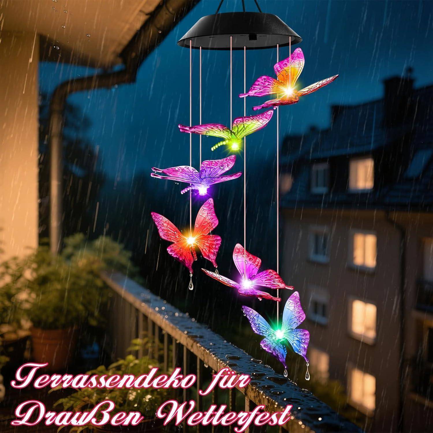 Ein bunter Schmetterlingswindspiel hängt an einem Balkon im Regen mit Stadtgebäuden im Hintergrund. Text lautet: 'Terrassendeko für Draußen Wetterfest'.