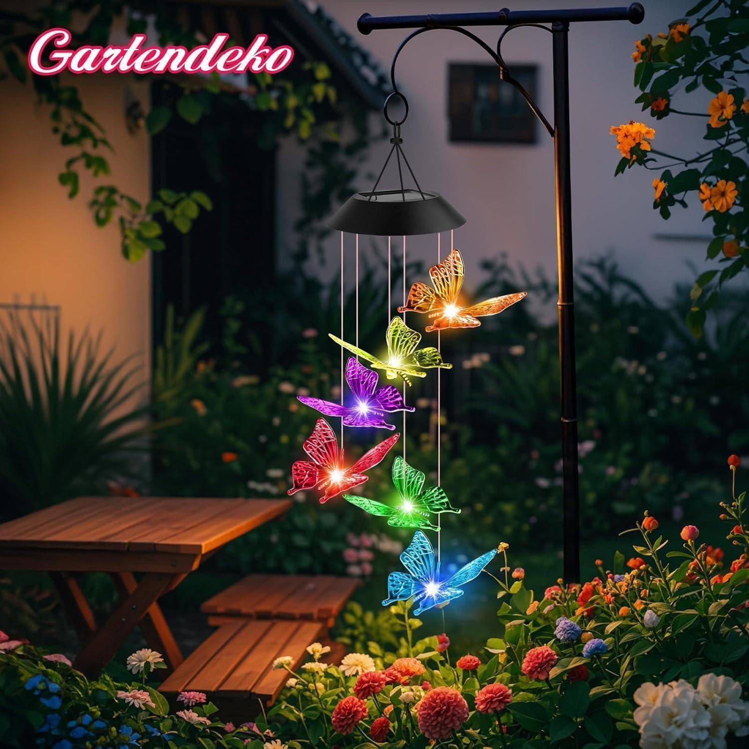 Eine Gartenszene in der Abenddämmerung mit einem dekorativen Windspiel aus bunten, beleuchteten Schmetterlingen, das neben einem Holzpicknicktisch hängt.