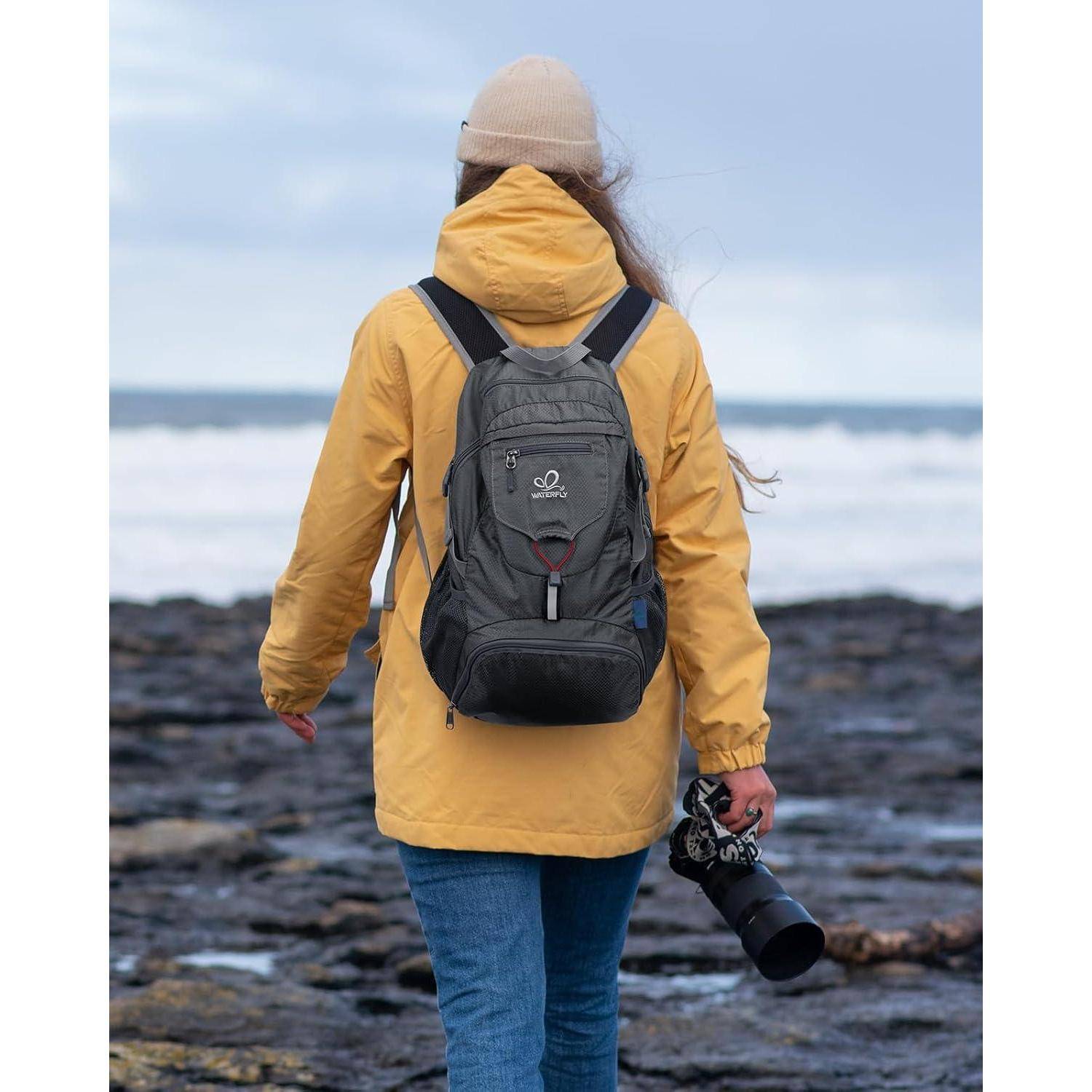 Eine Person in einer gelben Jacke und Mütze läuft an einem felsigen Strand entlang und hält eine Kamera in der einen Hand, während der Ozean im Hintergrund sichtbar ist.