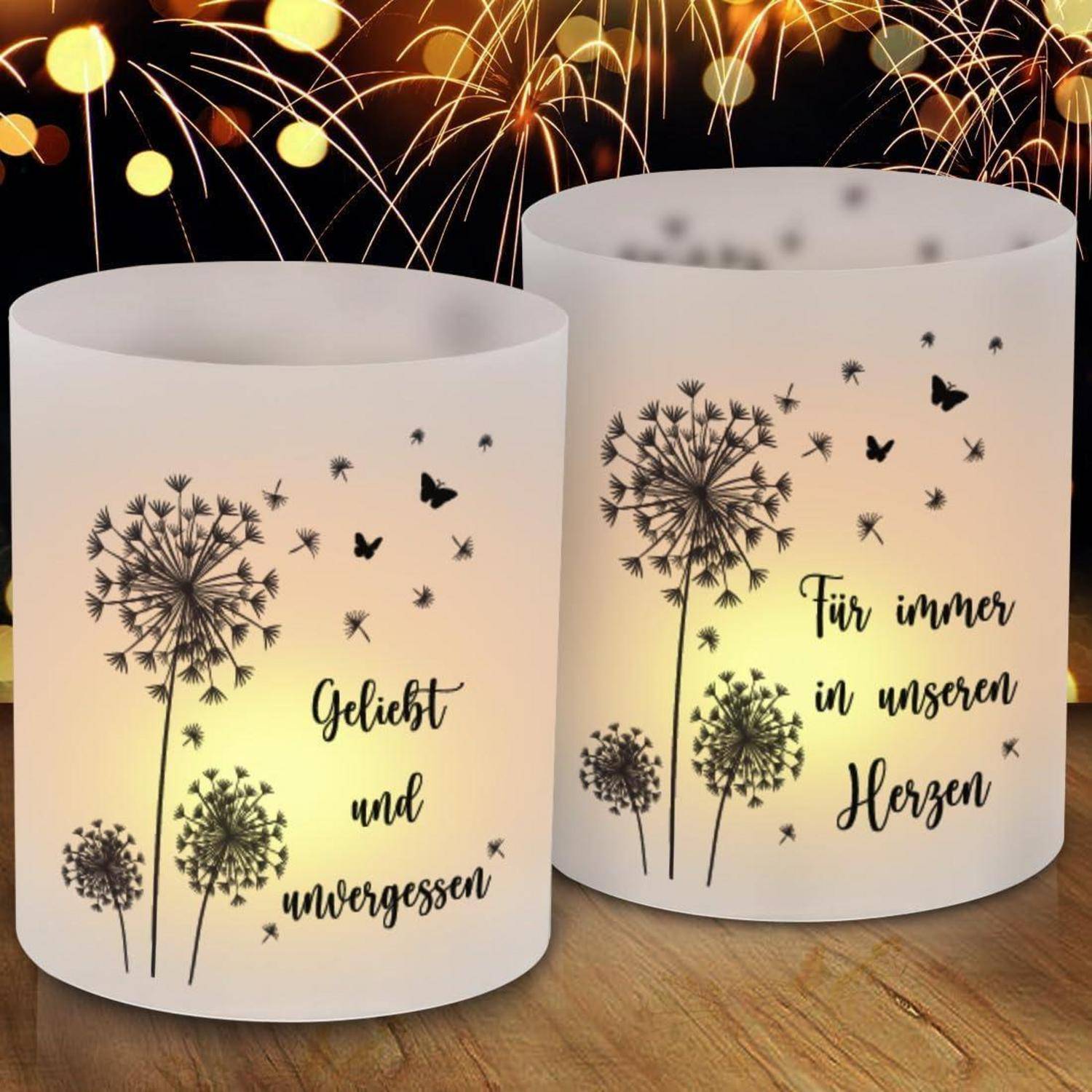 Zwei Kerzenhalter mit Löwenzahndesigns und deutschem Text: 'Geliebt und unvergessen' und 'Für immer in unseren Herzen' vor einem von Feuerwerk erleuchteten Himmel.