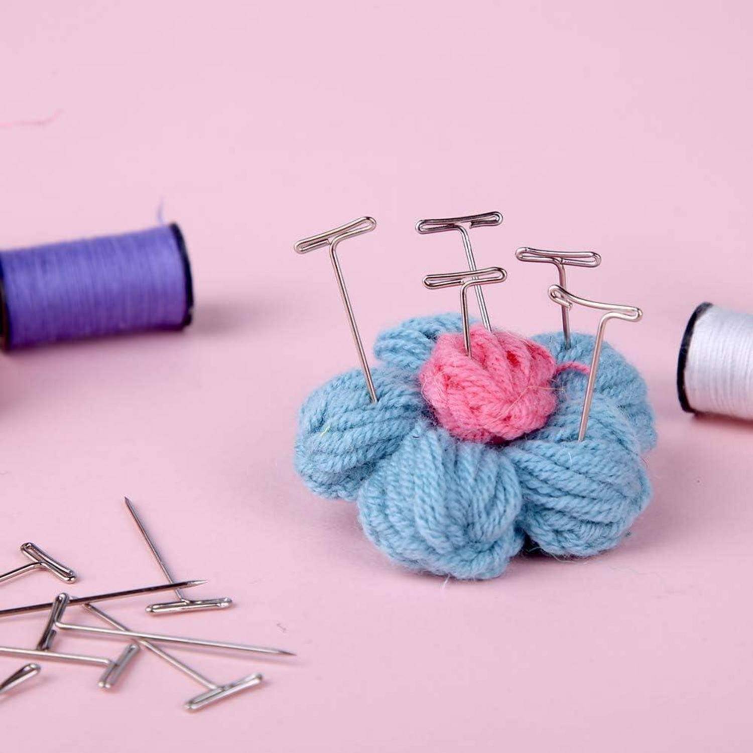 Eine kleine gestrickte Blume in Rosa und Blau mit T-förmigen Nadeln, umgeben von Garnrollen und Nähnadeln auf einem rosa Hintergrund.