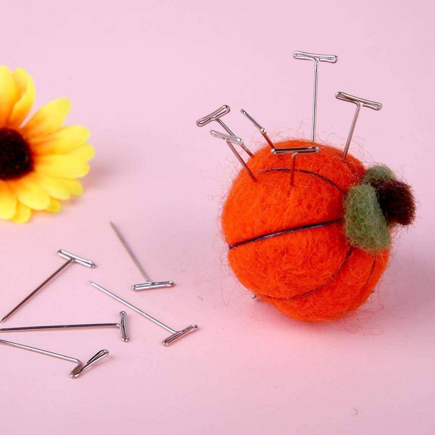 Eine Nadelkissen in Form eines Kürbisses mit T-Kopfstecknadeln, die daneben auf einer rosa Oberfläche verstreut liegen, neben einer gelben Gänseblümchen.
