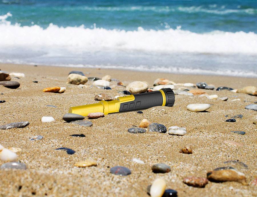 Gelb-schwarze Taschenlampe, die auf einem sandigen Strand zwischen Kieselsteinen liegt, mit Meereswellen im Hintergrund.