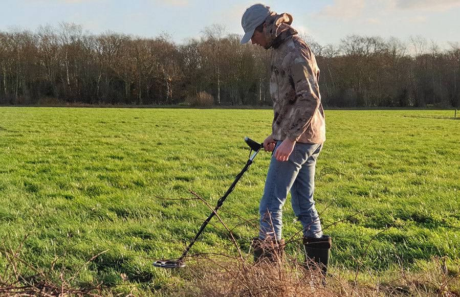 Person mit einem Metalldetektor auf einem grasbewachsenen Feld in der Nähe einer Baumreihe, der den Boden im Freien absucht.