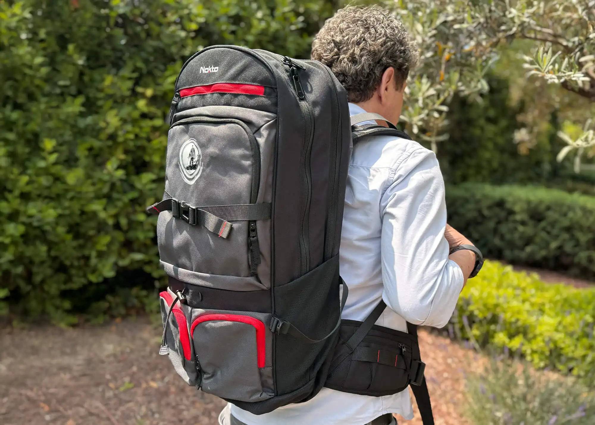 Person beim Wandern im Freien mit einem übergroßen schwarzen Rucksack auf dem Rücken in einer Garten- oder Wanderwegsumgebung.