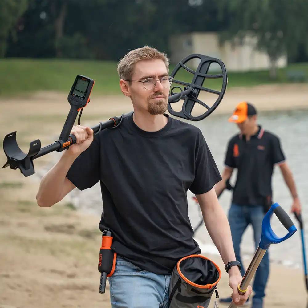 Mann mit einem Metalldetektor, Schaufel und Grabwerkzeugen an einem Strand, was auf eine Schatzsuche hindeutet.
