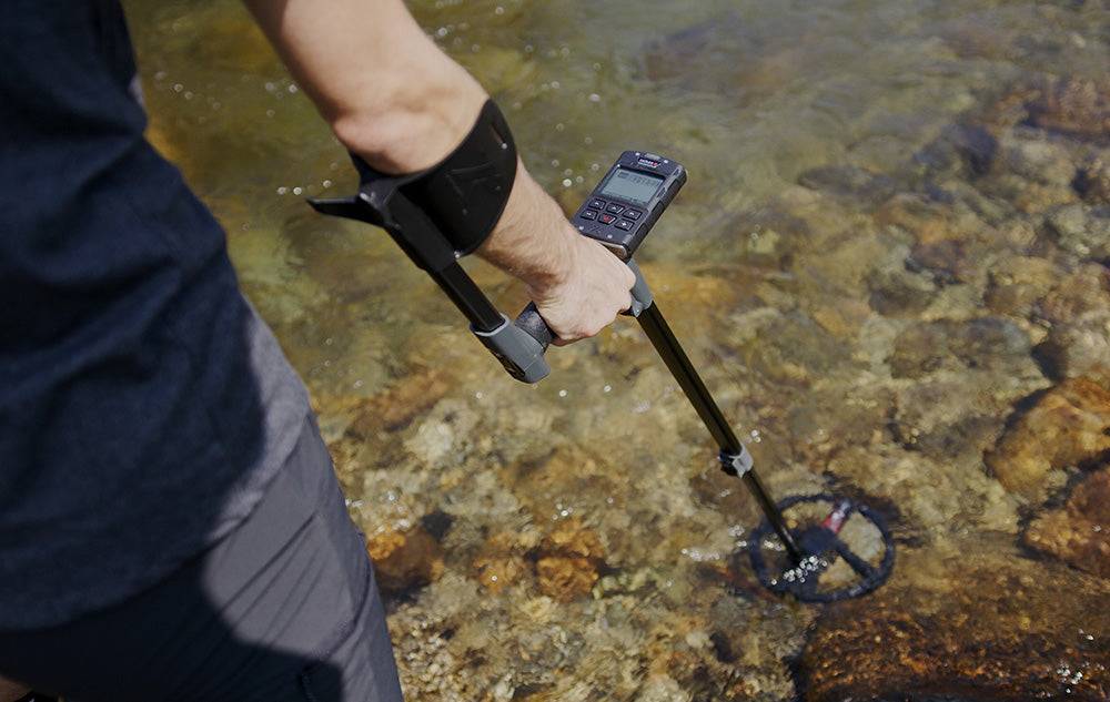 Eine Person verwendet einen Metalldetektor in der Nähe eines seichten Flusses oder Baches und tastet das Wasser und den felsigen Untergrund ab.