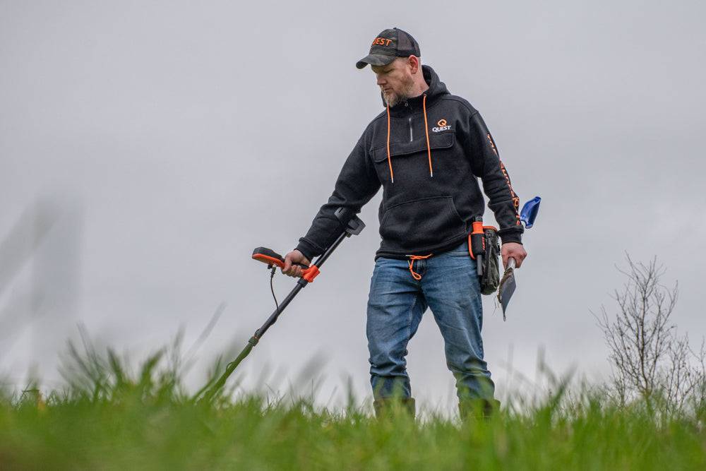 Ein Mann benutzt einen Metalldetektor auf einer grasbewachsenen Wiese und durchsucht den Boden im Freien an einem bewölkten Tag.