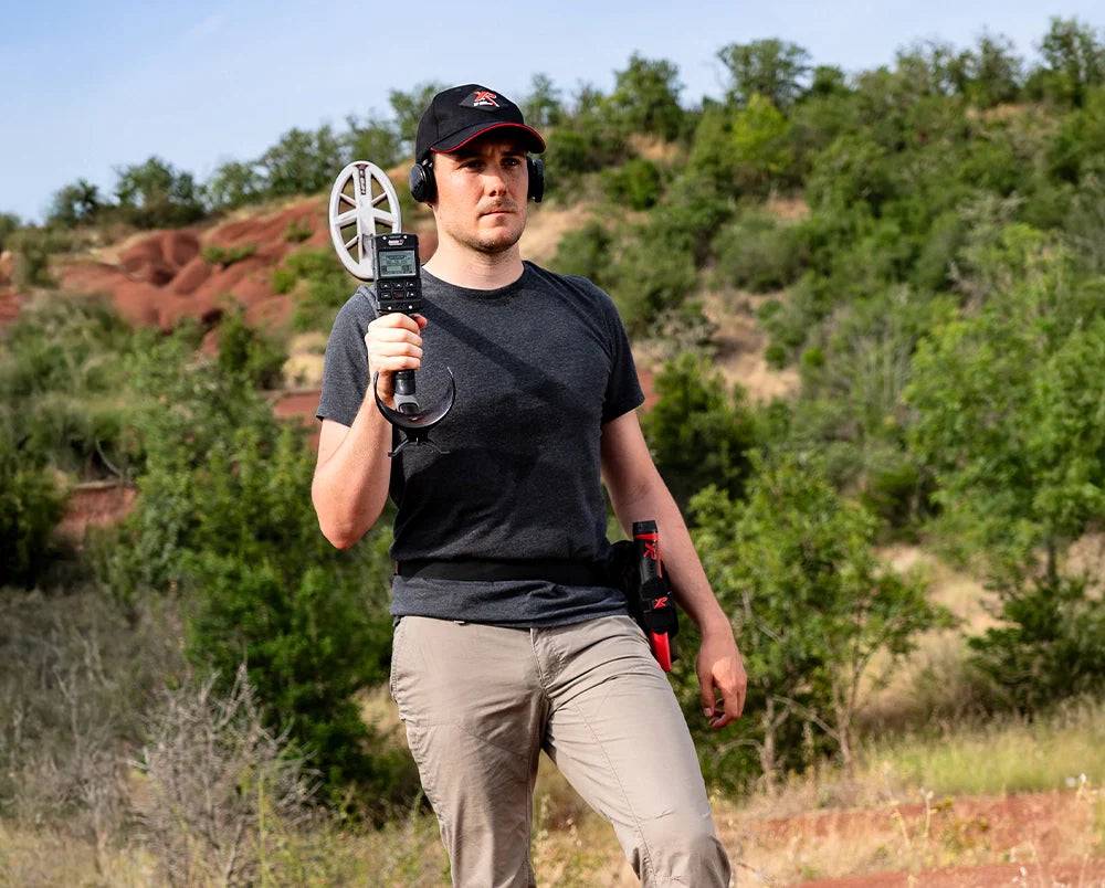 Eine Person mit einem Metallsuchgerät und einem GPS-Gerät, die durch ein grasiges, hügeliges Gebiet mit Bäumen im Hintergrund läuft und lässige Outdoor-Kleidung trägt.