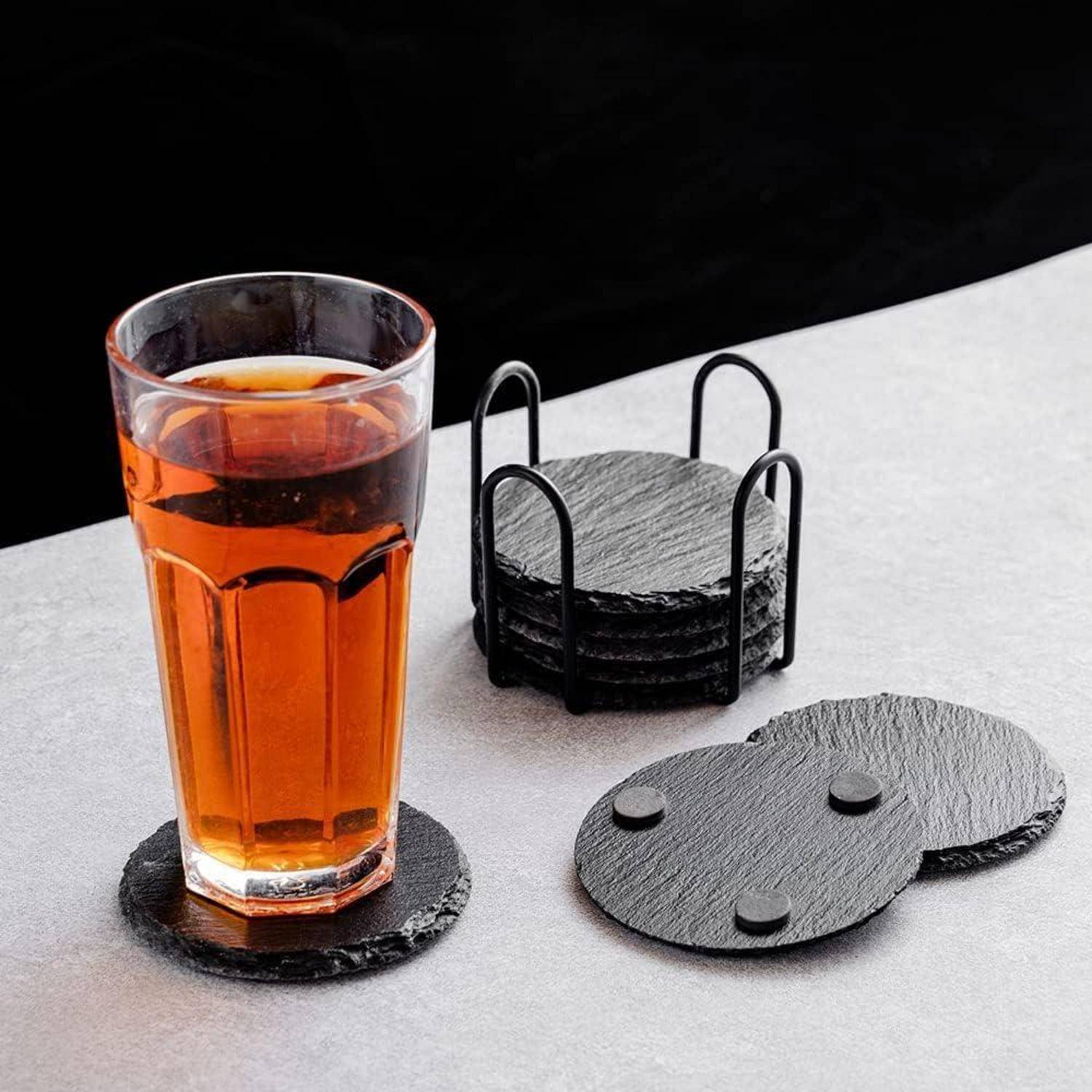 Ein Glas mit bernsteinfarbener Flüssigkeit auf einem runden schwarzen Schieferuntersetzer, mit einem Stapel ähnlicher Untersetzer und einem Halter im Hintergrund.