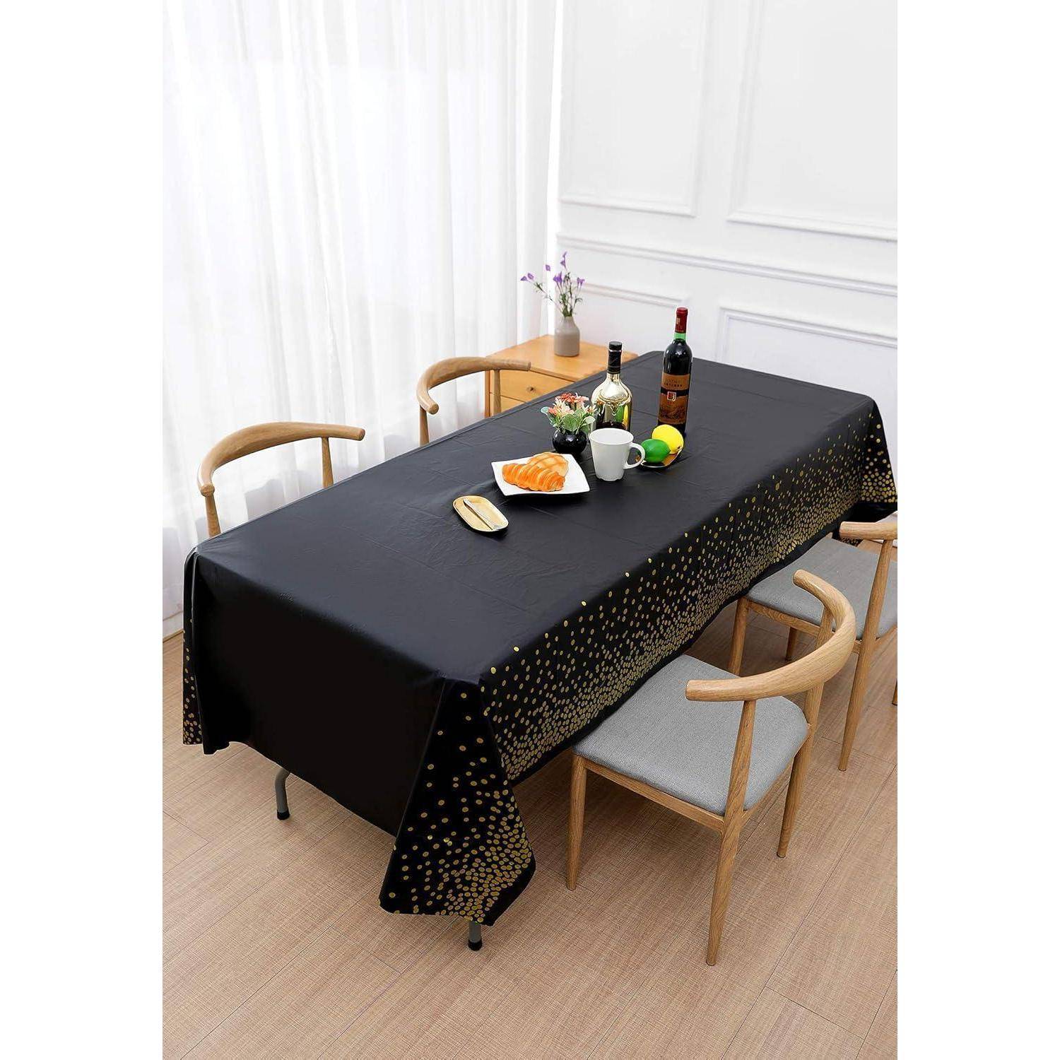 Ein Esstisch, der mit einer schwarzen Tischdecke mit goldenen Punkten ausgestattet ist, umgeben von sechs Holzstühlen. Auf dem Tisch befinden sich Teller, Besteck, Blumen und Weinflaschen.