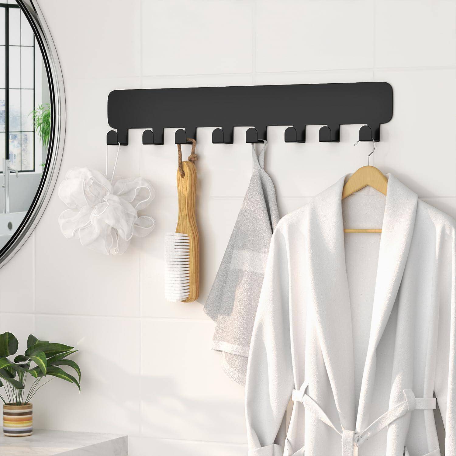 Ein weißer Bademantel hängt an einem modernen schwarzen Wandhaken-Ständer mit mehreren Haken in einem Badezimmer. In der Nähe befinden sich eine Pflanze und Badzubehör.